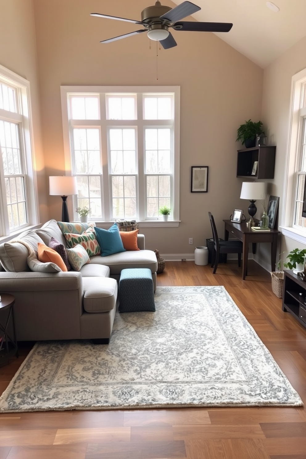 A cozy family room designed for comfort and functionality. It features a plush sectional sofa in a soft gray fabric, complemented by colorful throw pillows and a large area rug that adds warmth. In one corner, a small wooden desk is positioned for a work-from-home space, adorned with a stylish lamp and a few decorative items. The walls are painted in a light beige tone, and large windows allow natural light to flood the room, enhancing the inviting atmosphere.