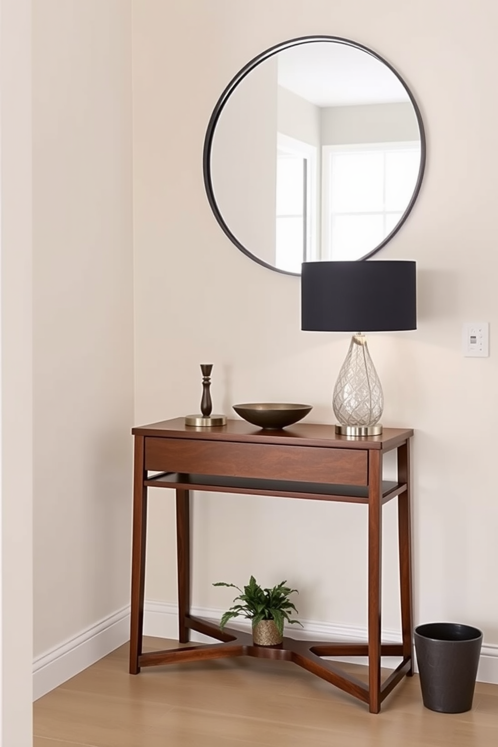 A narrow console table is positioned against the wall in a small foyer. The table features a sleek design with a rich wood finish and is adorned with a stylish lamp and a decorative bowl. Above the console table, a round mirror with a thin black frame reflects the light from the nearby window. The walls are painted in a soft cream color, and a small potted plant adds a touch of greenery to the space.