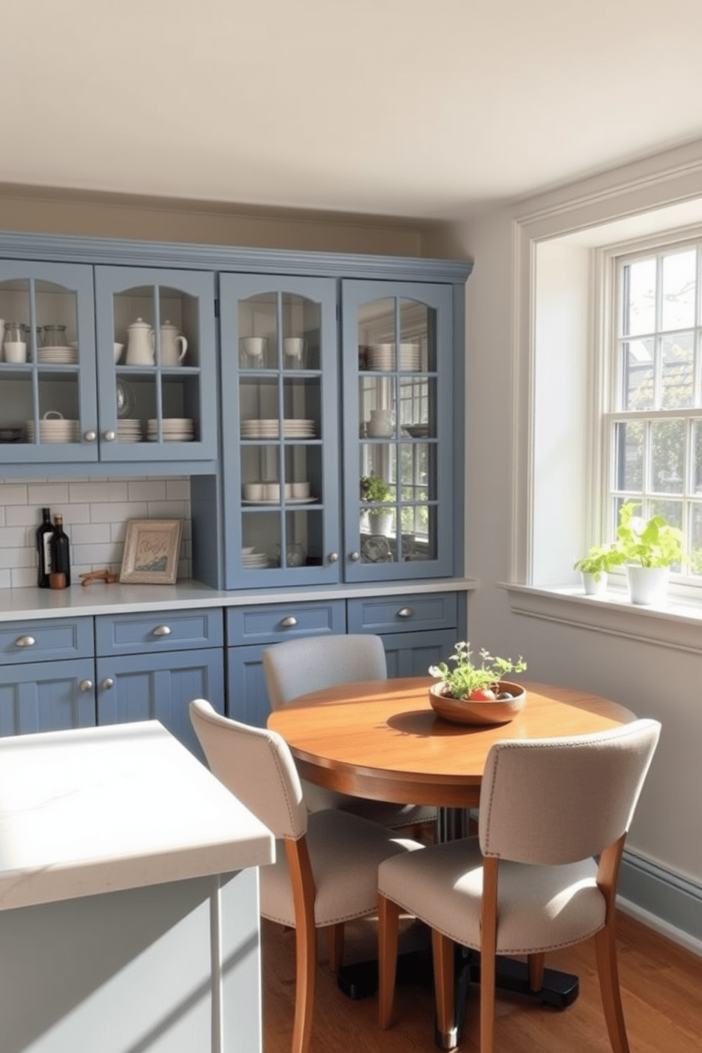 A cozy kitchen featuring glass cabinet doors that allow for easy visibility of dishware and decor. The cabinetry is painted in a soft blue hue, complementing the white subway tile backsplash and quartz countertops. A compact dining area with a round wooden table surrounded by upholstered chairs. Natural light floods the space through a large window, highlighting a small herb garden on the windowsill.