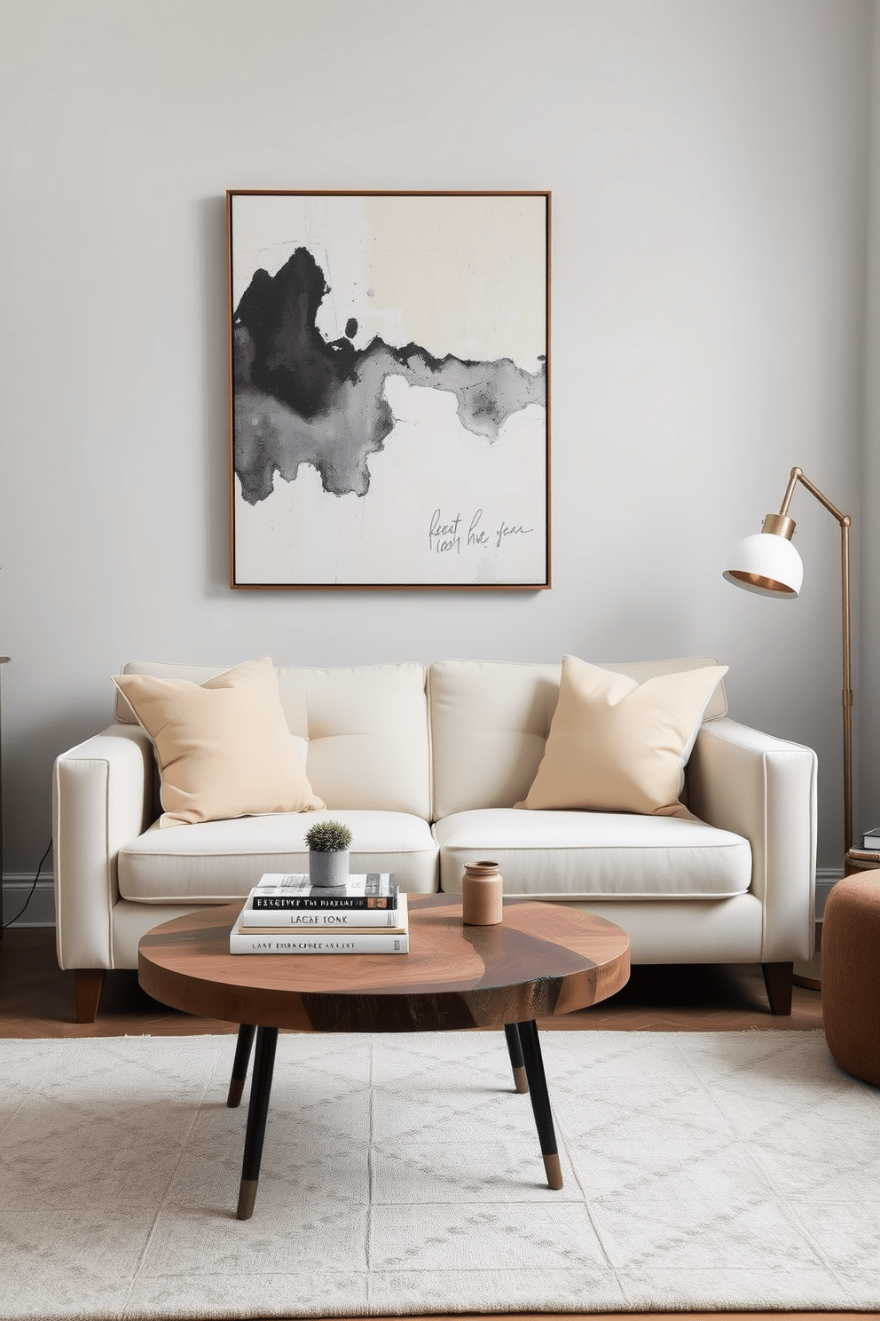 A cozy small living room with light gray walls and a plush cream-colored sofa positioned against one wall. A large piece of abstract artwork hangs high above the sofa, drawing the eye upward and creating an illusion of height. In front of the sofa, a round coffee table made of reclaimed wood is adorned with a stack of design books and a small potted plant. A soft area rug in muted tones anchors the seating area, while a floor lamp with a brass finish adds a touch of elegance.