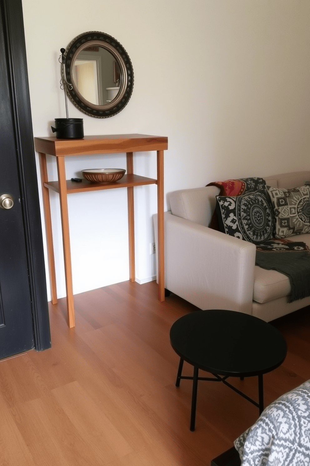 A cozy studio apartment featuring a small entryway console table made of reclaimed wood. The table is adorned with a stylish lamp and a decorative bowl, creating a warm welcome as you enter the space. The living area includes a compact sofa in a neutral fabric, paired with a colorful throw blanket and patterned cushions. A small round coffee table sits in front, complementing the overall aesthetic with its sleek design and inviting atmosphere.