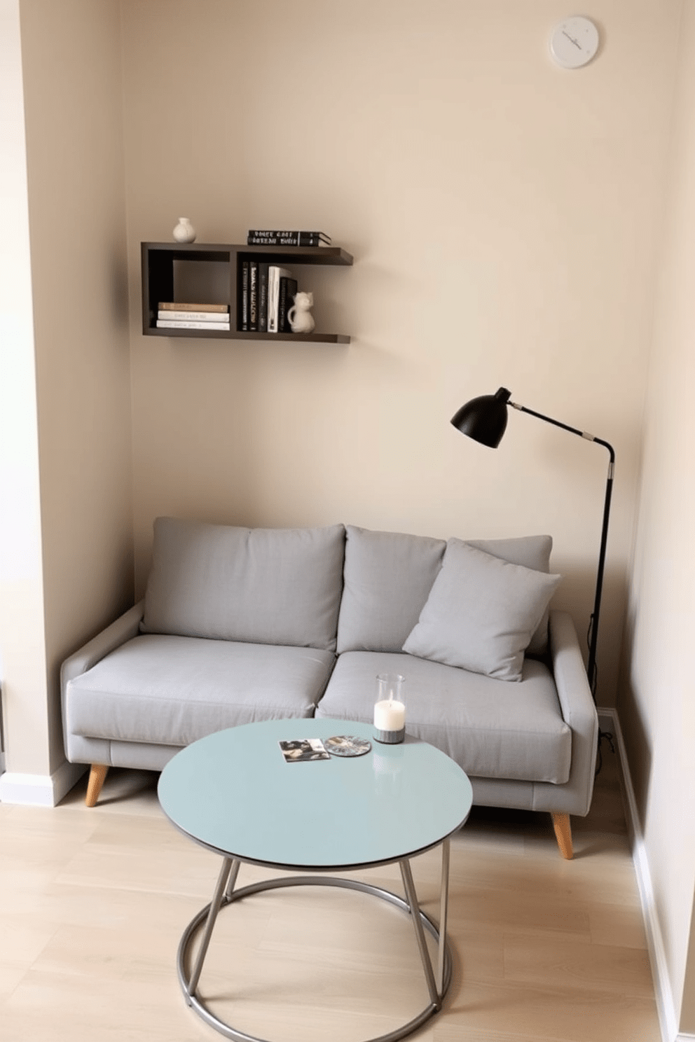 A compact sofa upholstered in soft grey fabric is placed against the wall, providing a cozy seating area. A small round coffee table with a glass top sits in front, adorned with a couple of stylish coasters and a decorative candle. To maximize space, a wall-mounted shelf above the sofa displays books and decorative items, adding personality to the room. The surrounding walls are painted in a light beige tone, enhancing the brightness and openness of the studio apartment.