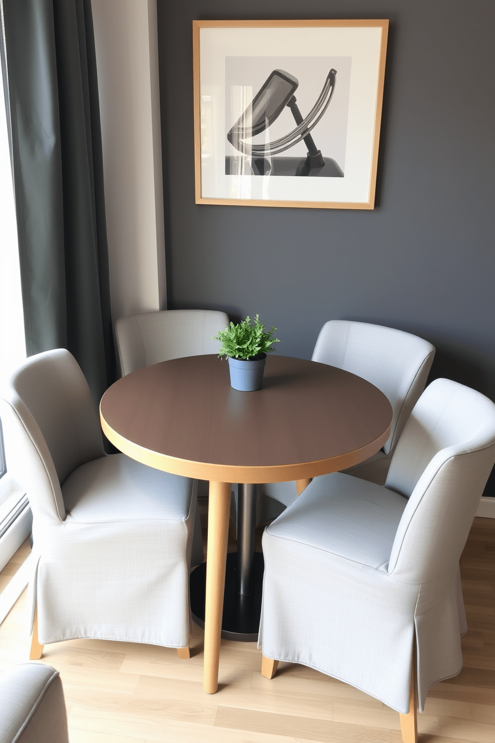 A compact dining table for meals in a studio apartment setting. The table is round with a light wood finish, surrounded by four upholstered chairs in a soft gray fabric. The dining area features a cozy nook by the window, with natural light streaming in. A small potted plant sits in the center of the table, adding a touch of greenery to the space.