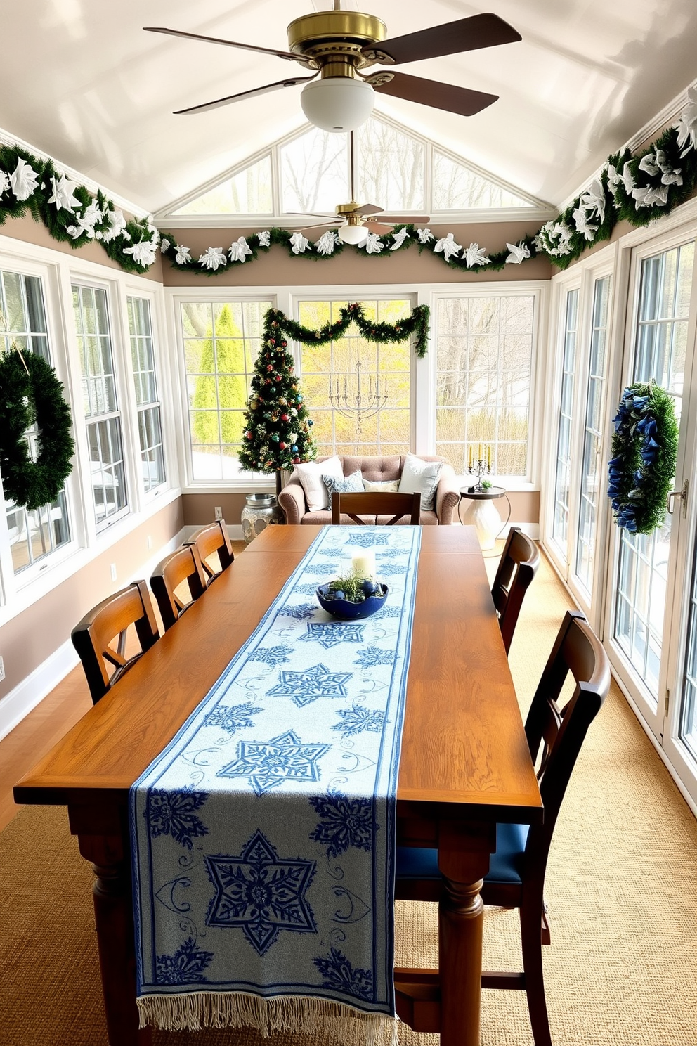 A sunroom decorated for Hanukkah features a long wooden dining table adorned with a beautiful holiday-themed table runner that showcases blue and silver motifs. Surrounding the table are comfortable chairs, and the sunroom is filled with natural light streaming through large windows, enhancing the festive atmosphere. On the walls, festive decorations such as blue and white garlands and menorahs are displayed, creating a warm and inviting space. A cozy seating area with plush cushions and a small coffee table is set up in one corner, perfect for enjoying holiday treats and gatherings.