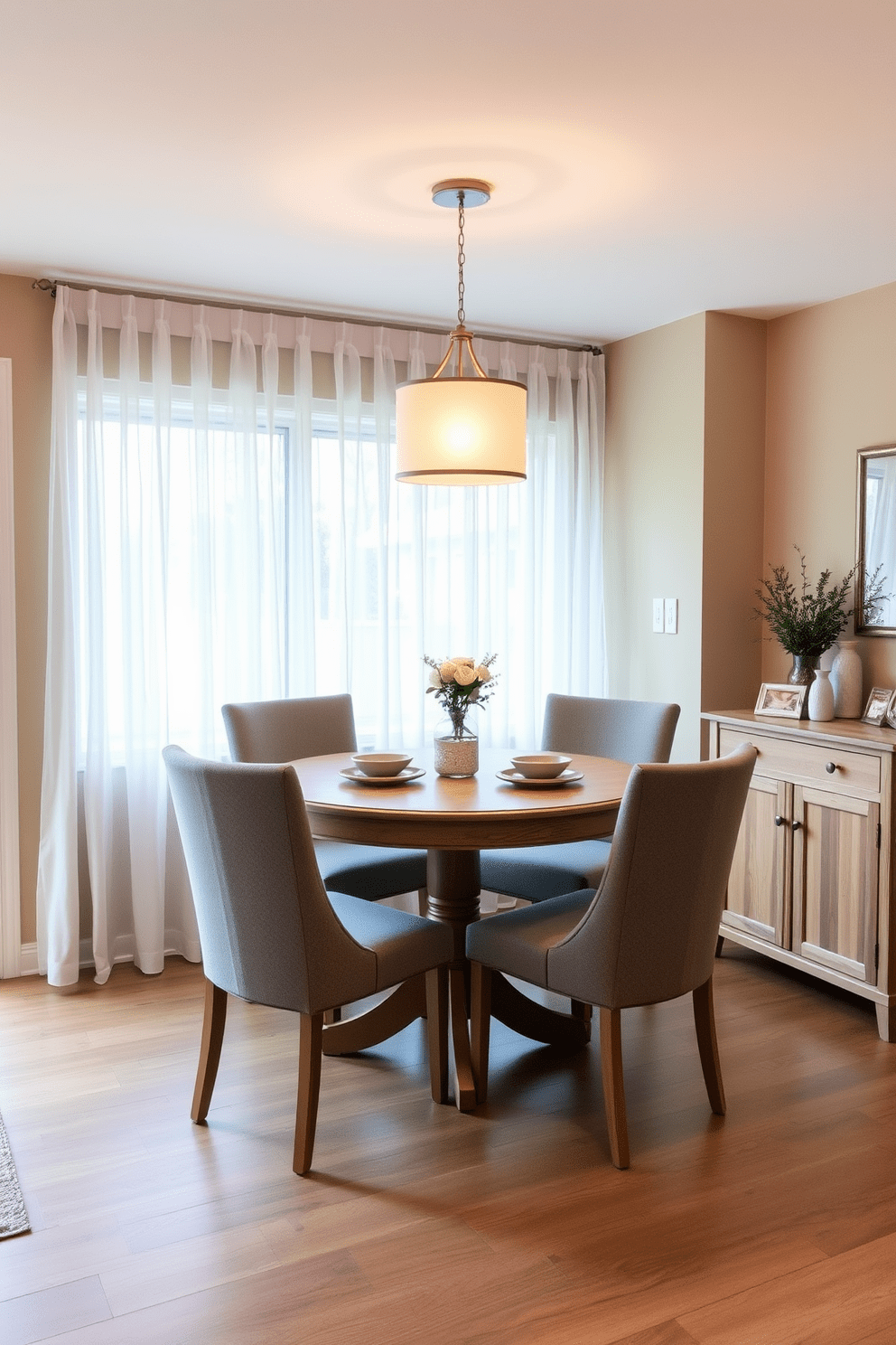 A cozy dining area featuring a small-scale round table with a light wood finish surrounded by four upholstered chairs in a soft gray fabric. The walls are painted in a warm beige tone, and a pendant light hangs above the table, creating an inviting atmosphere. To the side, there is a compact sideboard in a matching wood finish, adorned with decorative items and a small potted plant. A large window allows natural light to flood the space, with sheer curtains that add a touch of elegance while maintaining privacy.