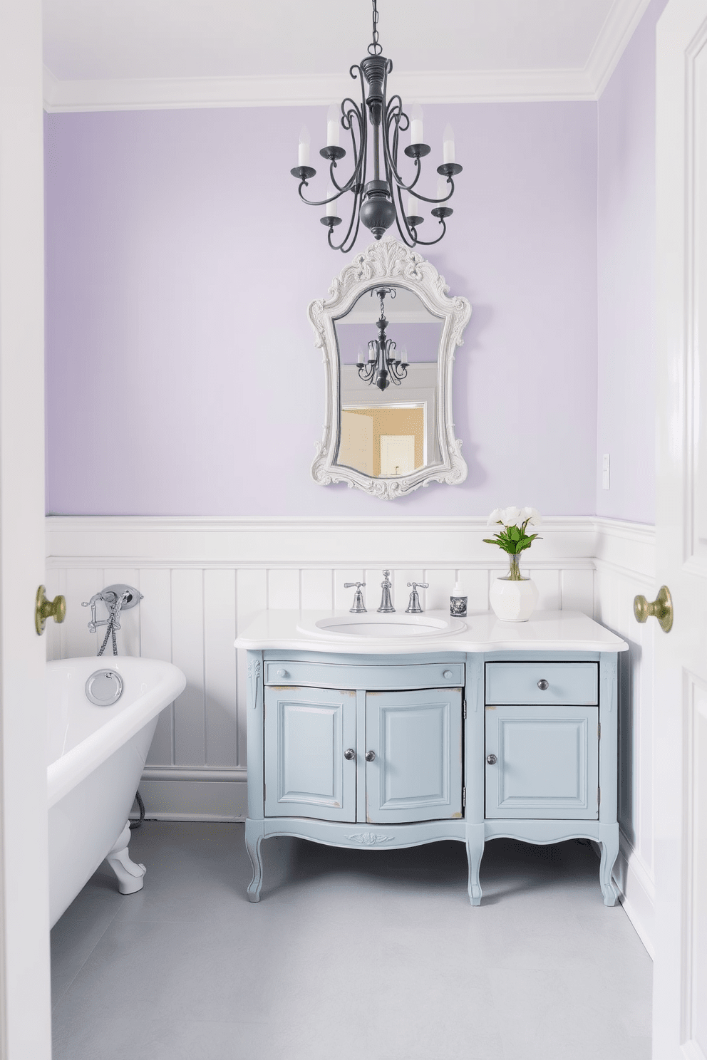 A serene traditional bathroom design featuring a soft pastel color palette. The walls are painted in a light lavender hue, complemented by white wainscoting and a vintage clawfoot bathtub. The floor is adorned with pale gray hexagonal tiles, while a wooden vanity with a soft blue finish holds a white porcelain sink. Above the vanity, an ornate mirror with a distressed white frame reflects the gentle lighting from a classic chandelier.