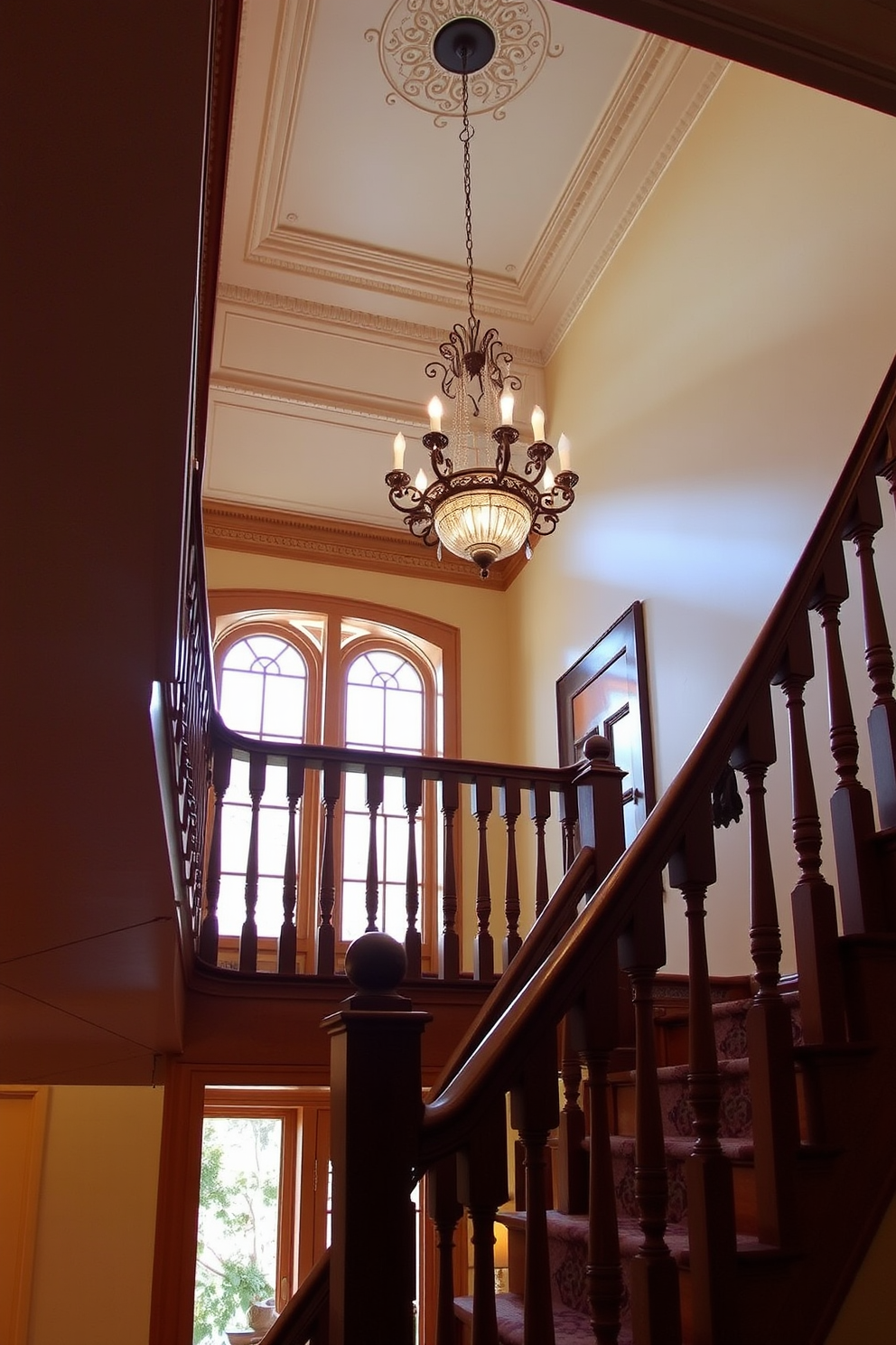 A traditional staircase features elegant wooden railings and balusters, leading to an upper landing adorned with intricate moldings. A grand chandelier hangs from the ceiling, casting a warm glow over the staircase and enhancing the classic charm of the space.