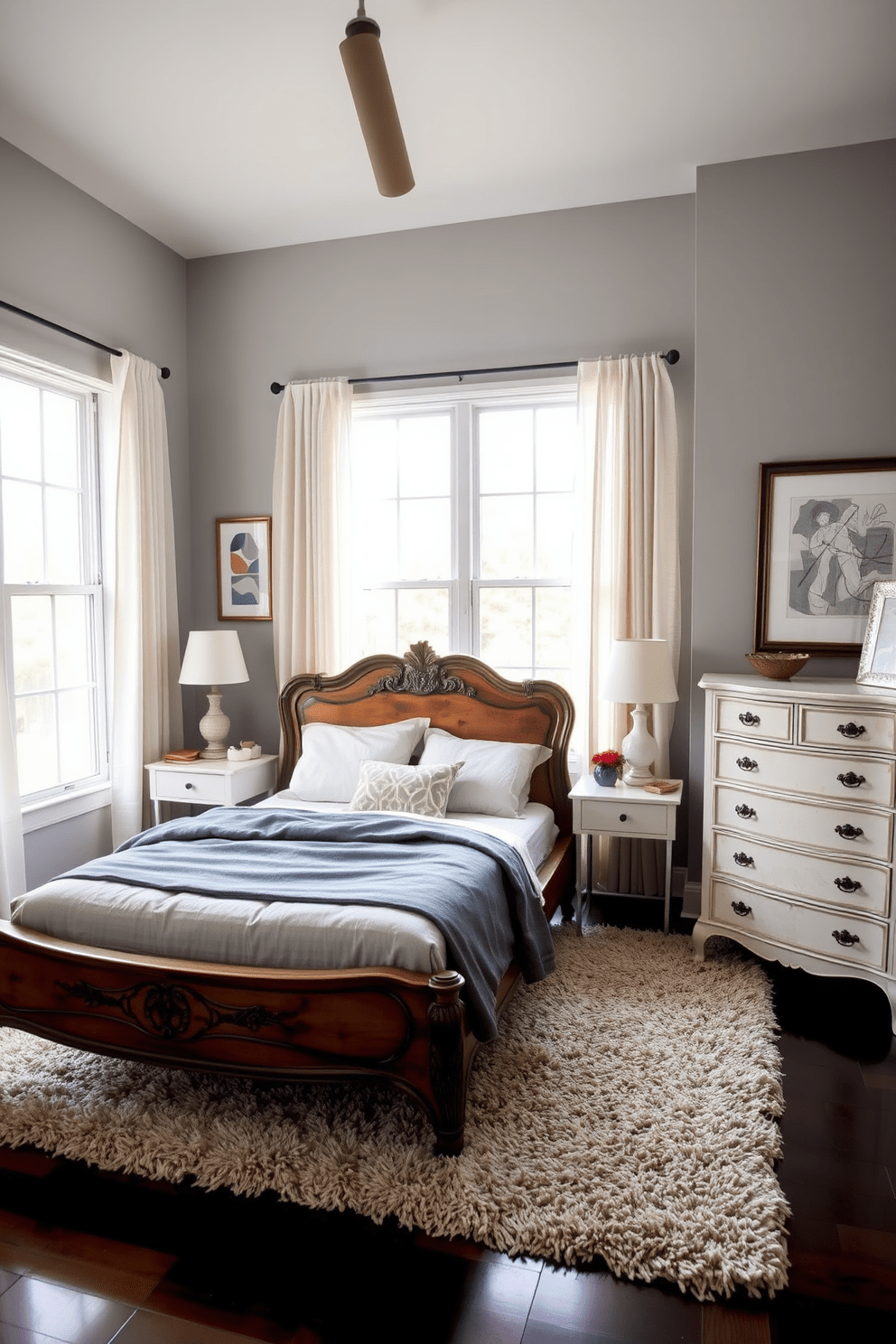 A cozy transitional bedroom featuring a vintage wooden bed frame with intricate carvings paired with sleek modern bedside tables. The walls are painted in a soft gray hue, and a plush area rug in neutral tones lies beneath the bed, adding warmth to the space. An antique dresser stands against one wall, complemented by contemporary artwork above it. Large windows dressed with sheer curtains allow natural light to flood the room, creating an inviting atmosphere.