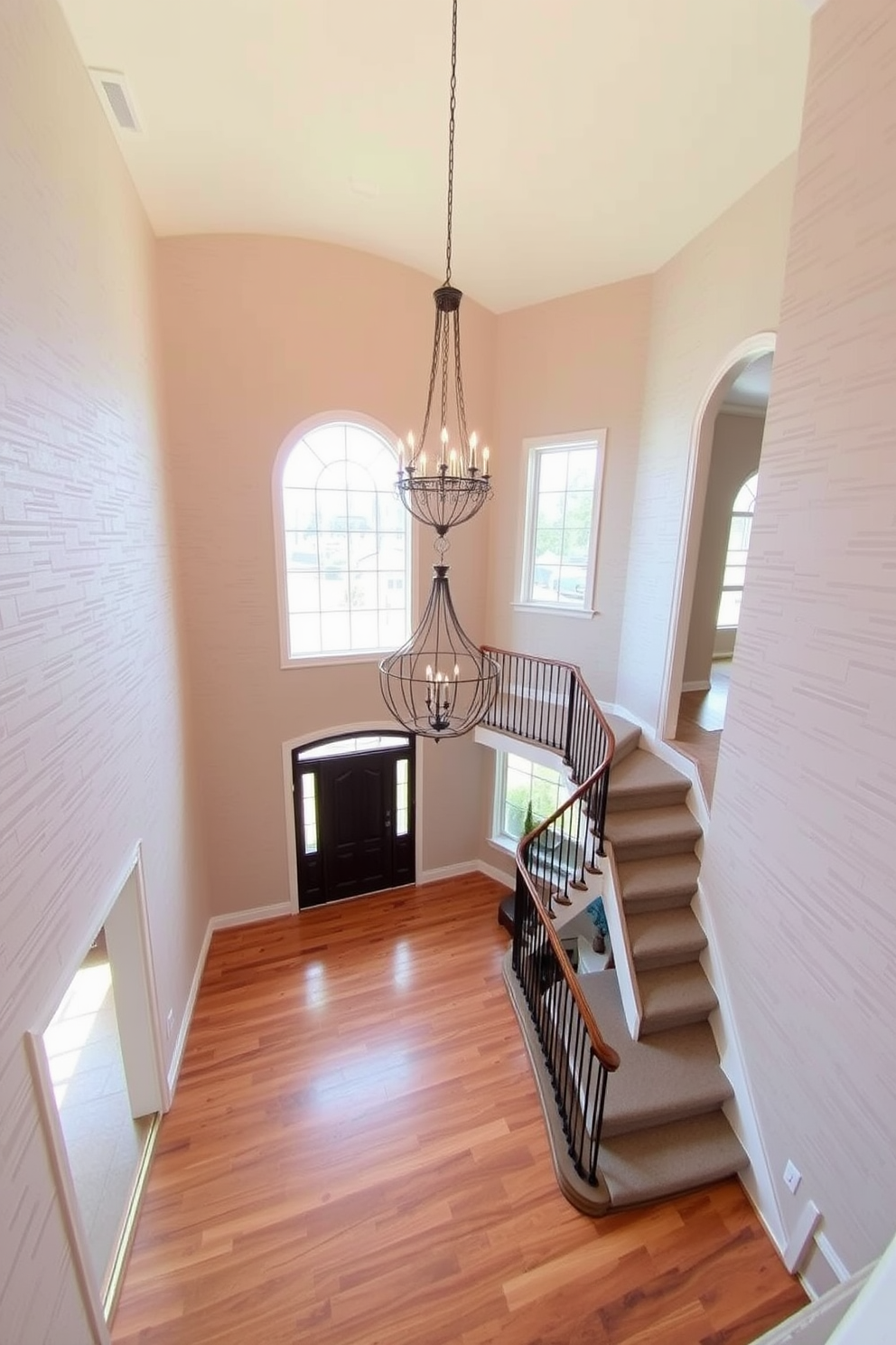 Textured wallpaper in a soft neutral tone covers the walls, adding depth and warmth to the space. A stunning chandelier hangs from the ceiling, casting a warm glow over the foyer. The two-story foyer features a grand staircase with elegant wrought iron railings. Large windows allow natural light to flood in, highlighting the beautiful hardwood floors below.