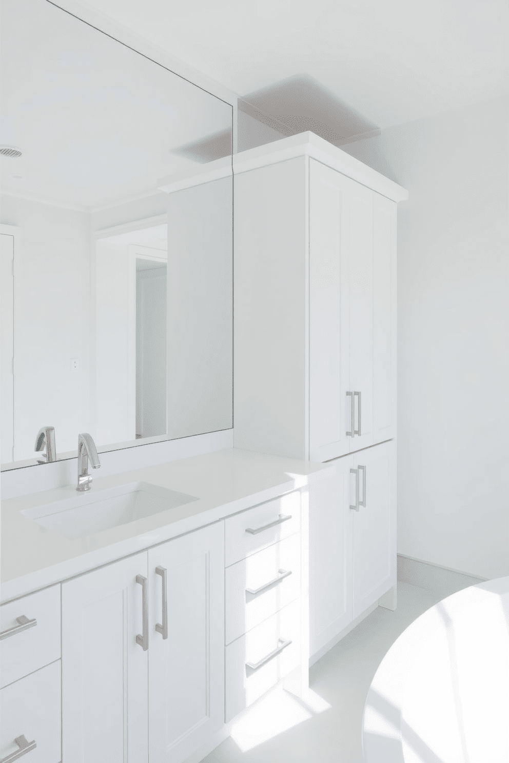 A minimalist white bathroom featuring cabinetry with sleek handles provides a clean and modern aesthetic. The space is enhanced by large mirrors and subtle lighting, creating an airy and open feel.