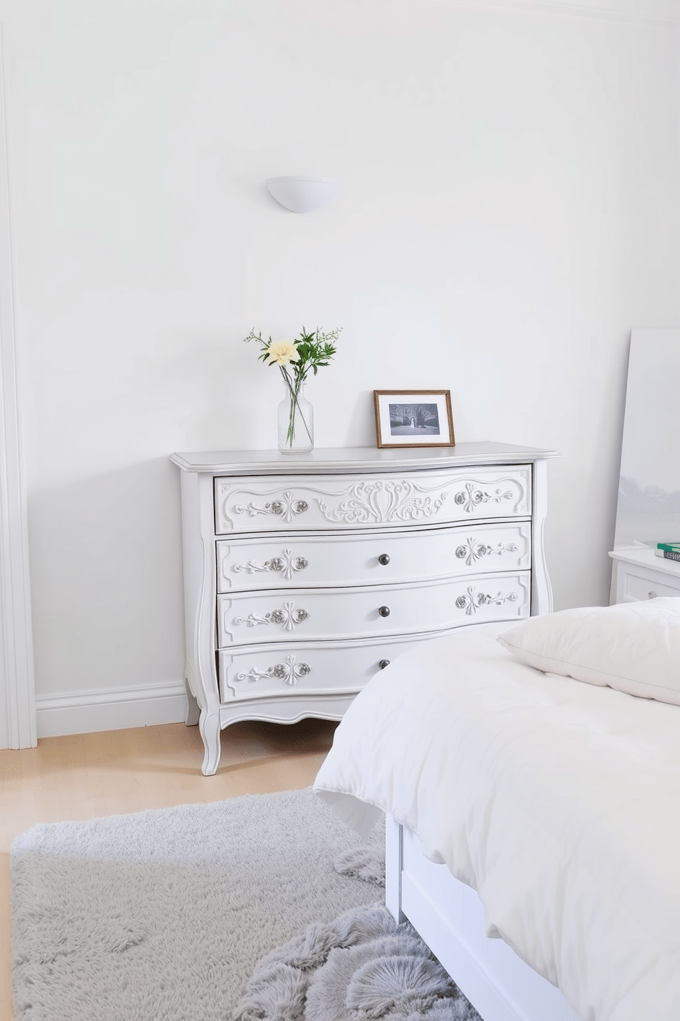 A vintage white dresser with intricate carvings stands against the wall, adding charm to the space. The dresser is adorned with a delicate vase of fresh flowers and a framed black-and-white photograph. The bedroom features soft white walls and a plush light gray area rug that complements the dresser. A cozy bed with fluffy white bedding and decorative pillows creates a serene and inviting atmosphere.