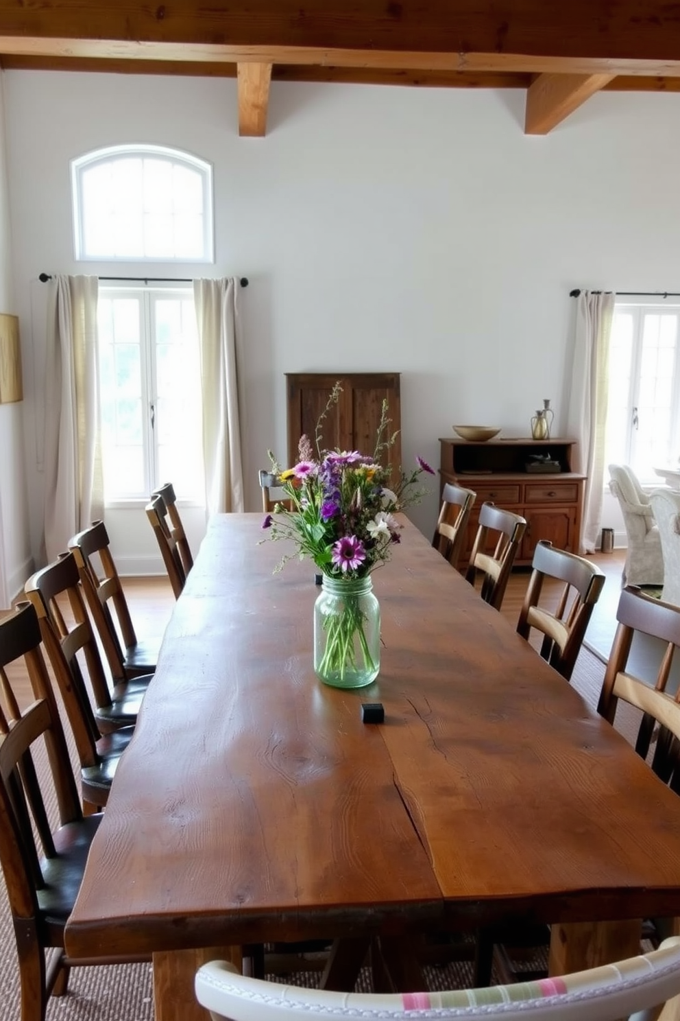 A rustic farmhouse dining room features a long reclaimed wood table surrounded by mismatched wooden chairs. The walls are painted in a soft white, with exposed wooden beams adding warmth to the space. Natural light floods the room through large windows adorned with simple linen curtains. A centerpiece of wildflowers in a mason jar sits on the table, complemented by vintage tableware.