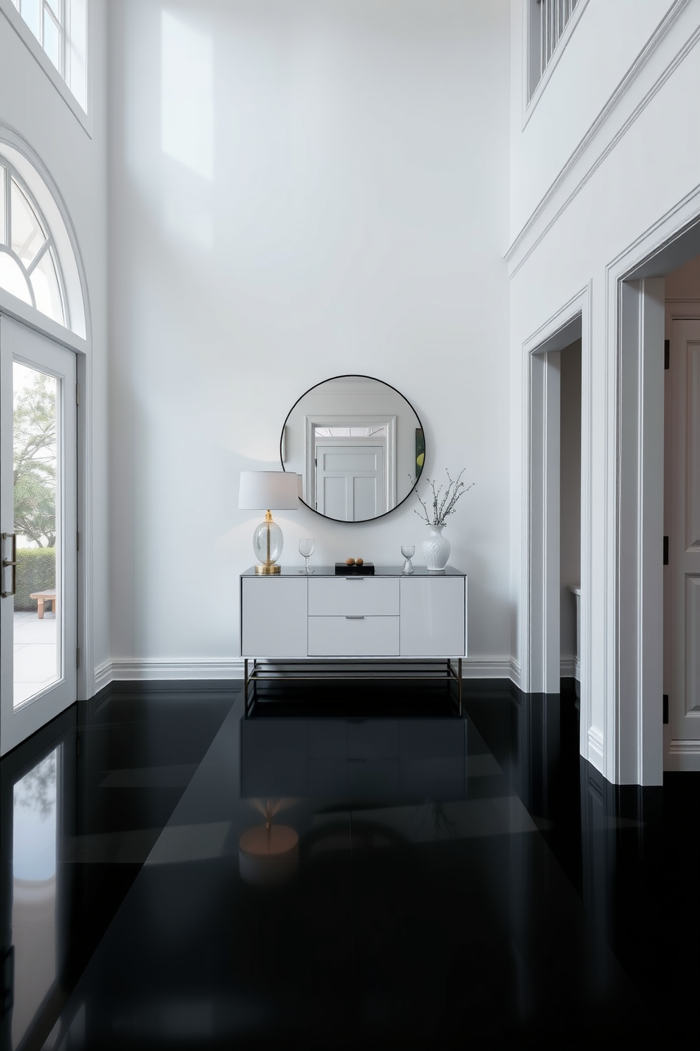 A bright white foyer with high ceilings creates an inviting atmosphere. The black flooring contrasts elegantly with the white walls, adding a modern touch. A sleek console table with a minimalist design sits against the wall, adorned with a stylish lamp and decorative items. A large round mirror hangs above the table, enhancing the sense of space and light in the foyer.