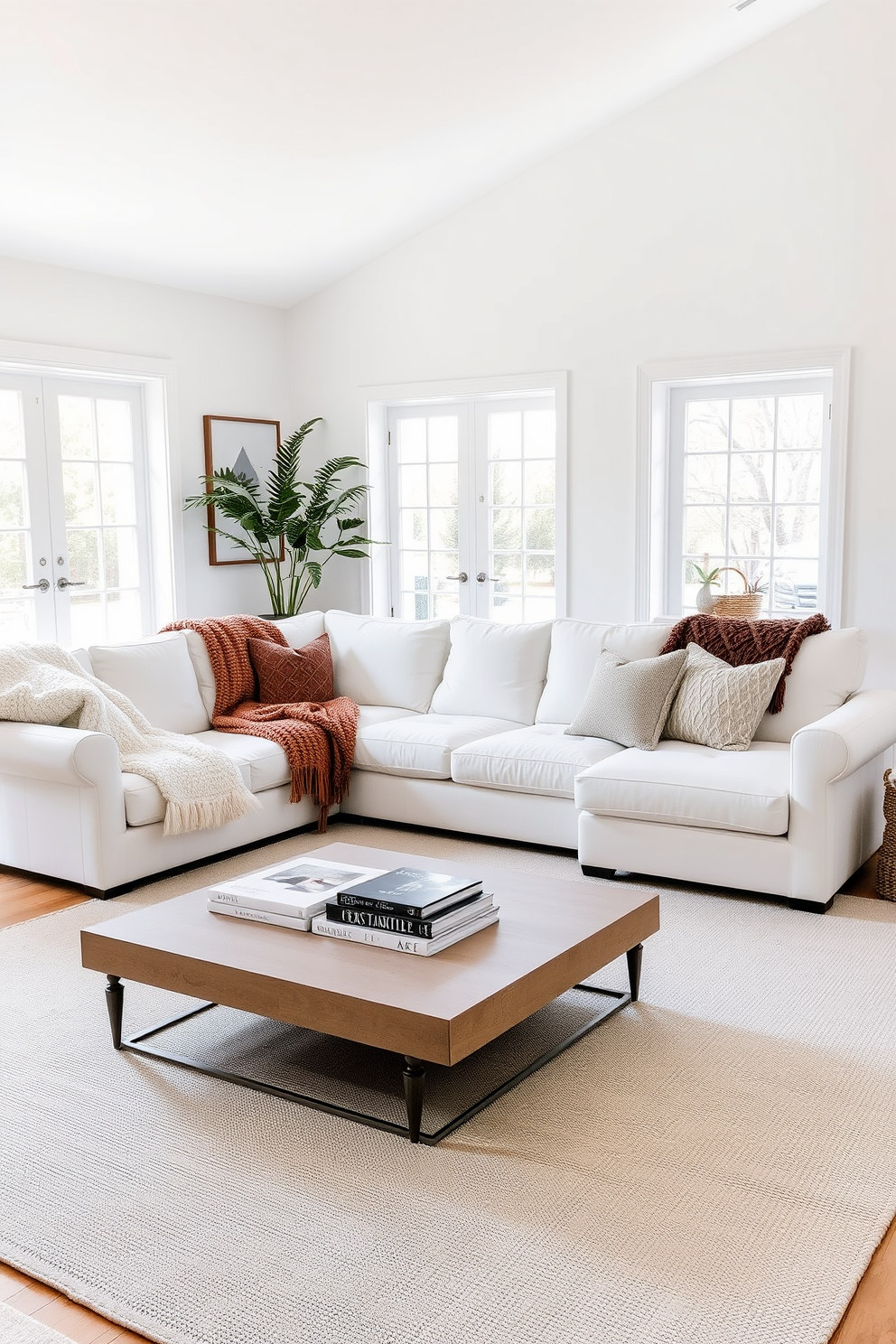 A bright and airy living room featuring a large white sectional sofa adorned with lush throw blankets in various textures and colors. The walls are painted in a soft white, and large windows allow natural light to flood the space, enhancing the cozy atmosphere. A minimalist coffee table sits in front of the sofa, topped with a few art books and a decorative tray. The floor is covered with a light gray area rug, adding warmth and comfort to the overall design.