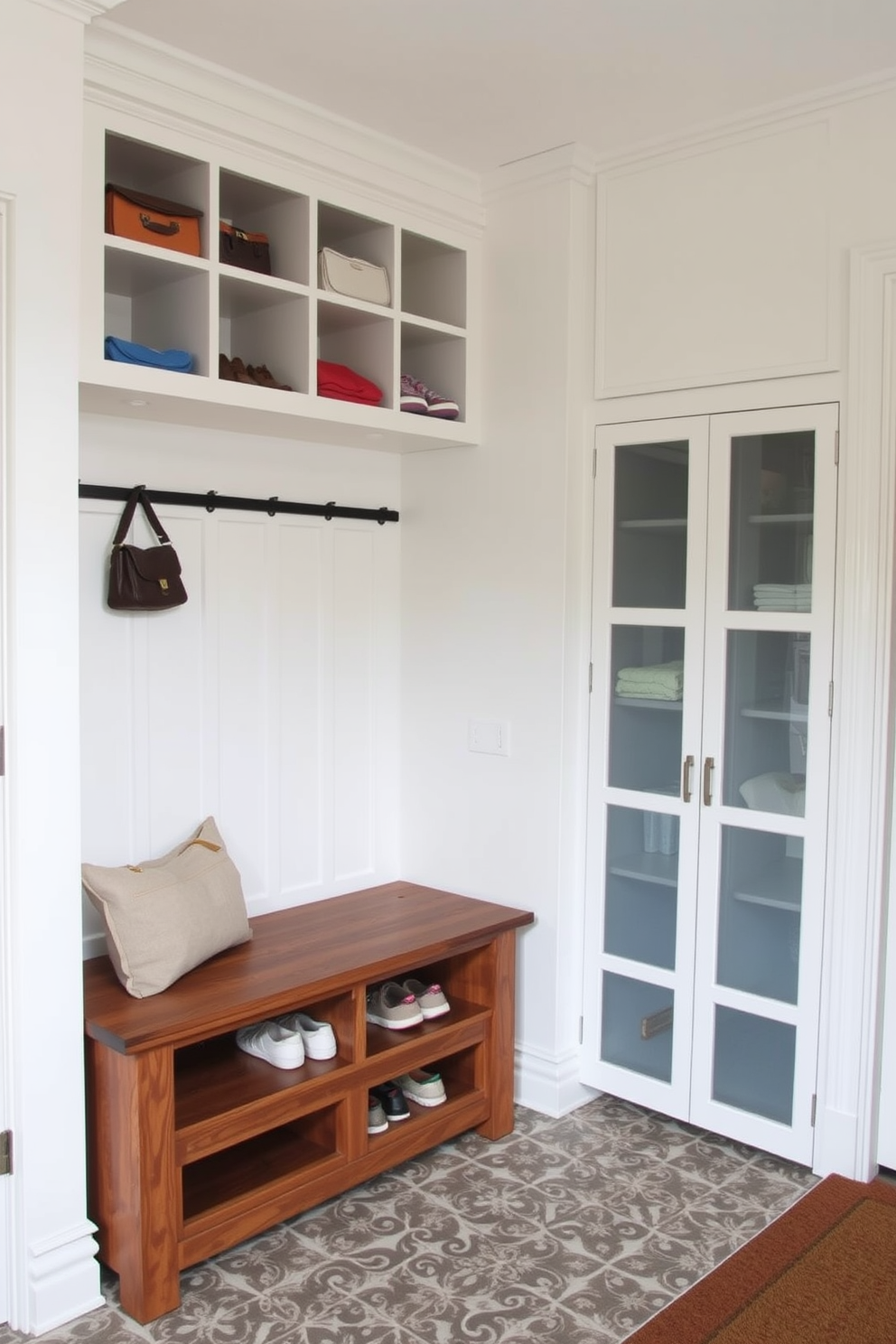 A stylish mudroom featuring a mix of open and closed storage solutions. The space includes a large wooden bench with built-in cubbies above for easy access to shoes and bags. To the side, a tall cabinet with frosted glass doors provides concealed storage for cleaning supplies and outdoor gear. The walls are painted in a soft white, complemented by a patterned tile floor that adds a touch of elegance.