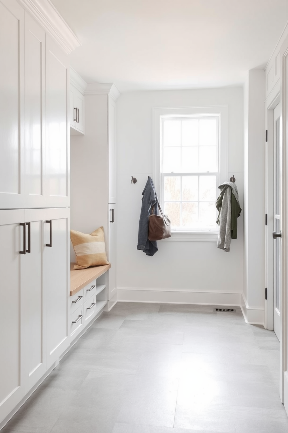 A minimalist mudroom designed for a modern feel features sleek white cabinetry with clean lines and ample storage. The floor is adorned with large light gray tiles, providing a spacious and airy atmosphere. A simple bench with a cushion sits against the wall, accompanied by hooks for hanging coats and bags. Natural light floods the space through a large window, enhancing the bright and uncluttered aesthetic.