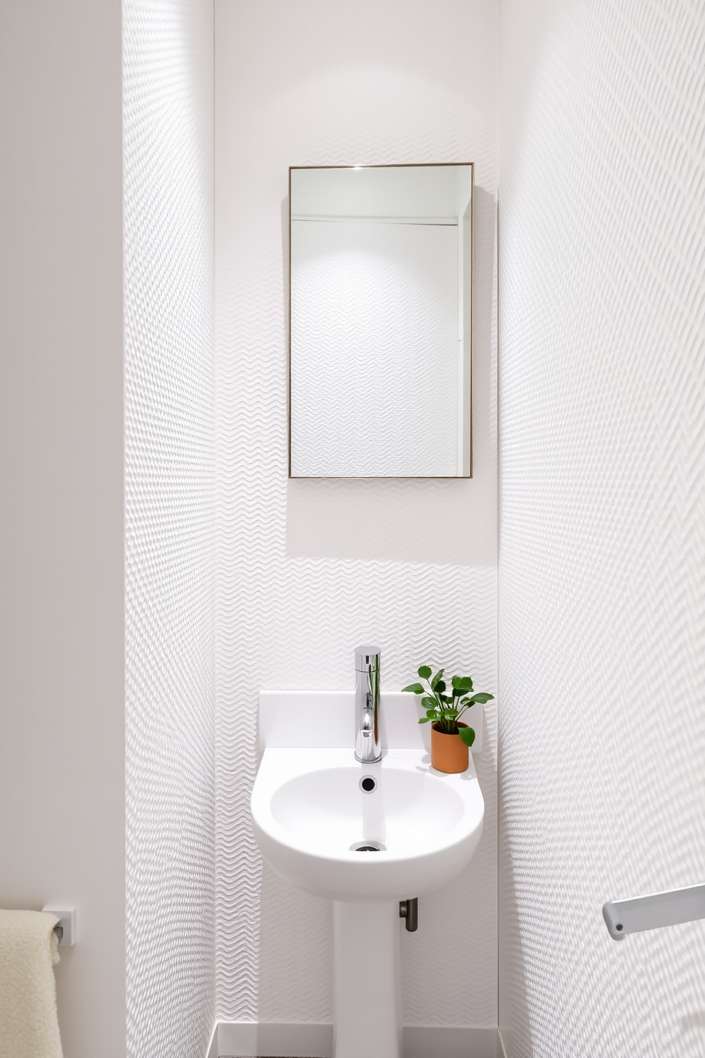 A compact corner sink is elegantly integrated into a stylish white powder room. The walls are adorned with textured white tiles, creating a bright and airy atmosphere. A sleek mirror with a minimalist frame hangs above the sink, reflecting the soft lighting. A small shelf beside the sink displays neatly rolled towels and a decorative plant, adding a touch of greenery.