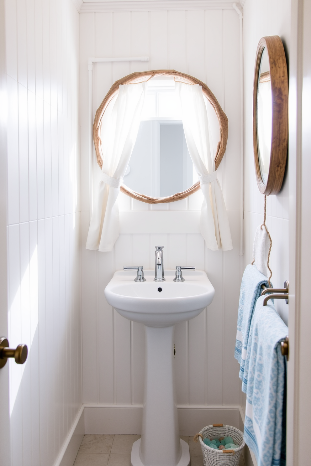 A bright white beadboard lines the walls of a charming coastal powder room. The space features a sleek white pedestal sink and a round mirror framed in driftwood, evoking a serene beach vibe. Natural light floods in through a small window adorned with sheer linen curtains. Soft blue accents are introduced through decorative towels and a small vase filled with fresh sea glass.