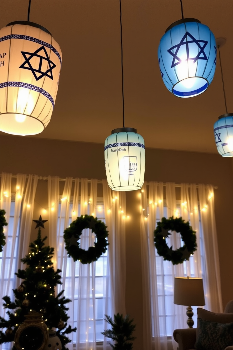 A cozy living room adorned with hanging paper lanterns featuring Hanukkah designs. The lanterns cast a warm glow, creating a festive atmosphere that complements the blue and silver color scheme of the decor. The windows are beautifully decorated with sheer white curtains that allow natural light to filter through. String lights are draped along the window frames, enhancing the holiday spirit and providing a charming backdrop for the season.