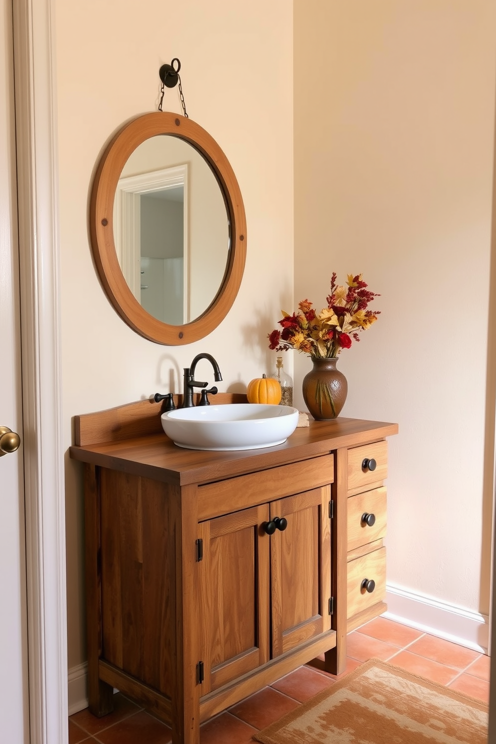 A cozy fall bathroom setting with warm tones. A wooden vanity with a rustic finish holds a large ceramic sink, and above it, a round mirror with a natural wood frame reflects the soft light. On the countertop, a vase filled with dried flowers in autumn hues adds a touch of seasonal charm. The walls are painted in a soft cream color, and the floor is adorned with warm-toned, textured tiles.