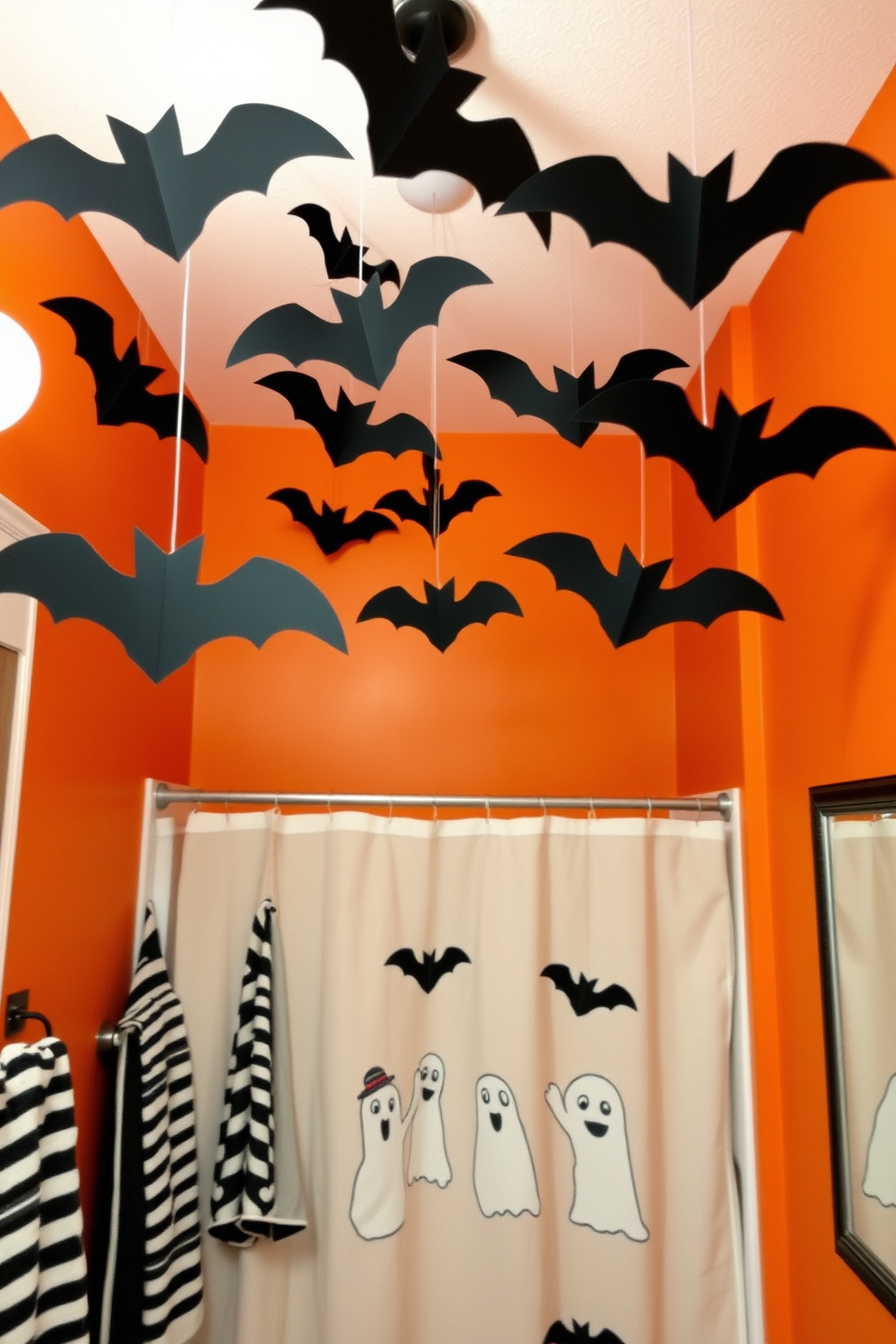 A whimsical Halloween bathroom setting. The ceiling is adorned with hanging bats made of black paper, creating a spooky atmosphere. The walls are painted in a deep orange hue, complemented by black and white striped towels. A ghostly shower curtain features playful ghost designs, adding to the festive decor.