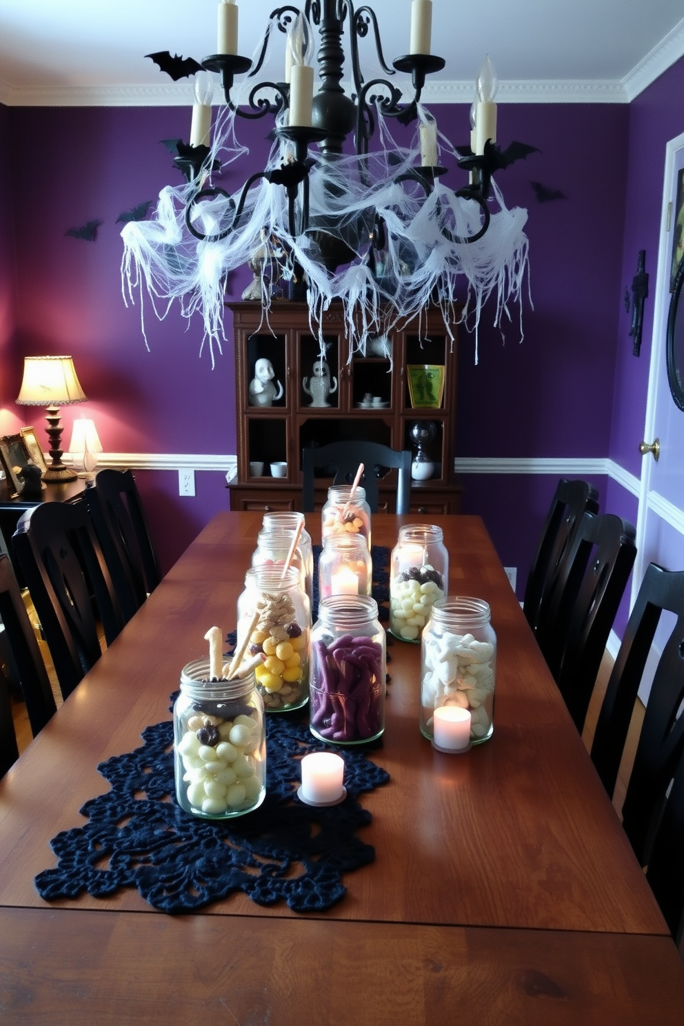 Eerie glass jars filled with assorted spooky treats are placed on a long wooden dining table. The table is adorned with a dark lace table runner, and flickering candlelight casts shadows that enhance the haunting atmosphere. Above the table, a vintage chandelier drips with cobwebs and small bats hang from its arms. The walls are painted in deep purple, and ghostly decorations are scattered throughout the room, creating an inviting yet chilling dining experience.