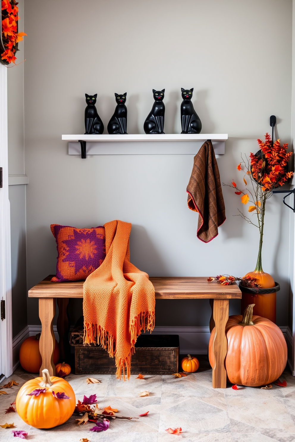 A cozy mudroom adorned for Halloween features a rustic bench with a warm throw blanket draped over it. Black cat figurines are playfully arranged on a shelf, adding a whimsical touch to the seasonal decor. The walls are painted in a soft gray, providing a neutral backdrop for vibrant orange and purple accents. Pumpkins and autumn leaves are scattered around, enhancing the festive atmosphere of the space.