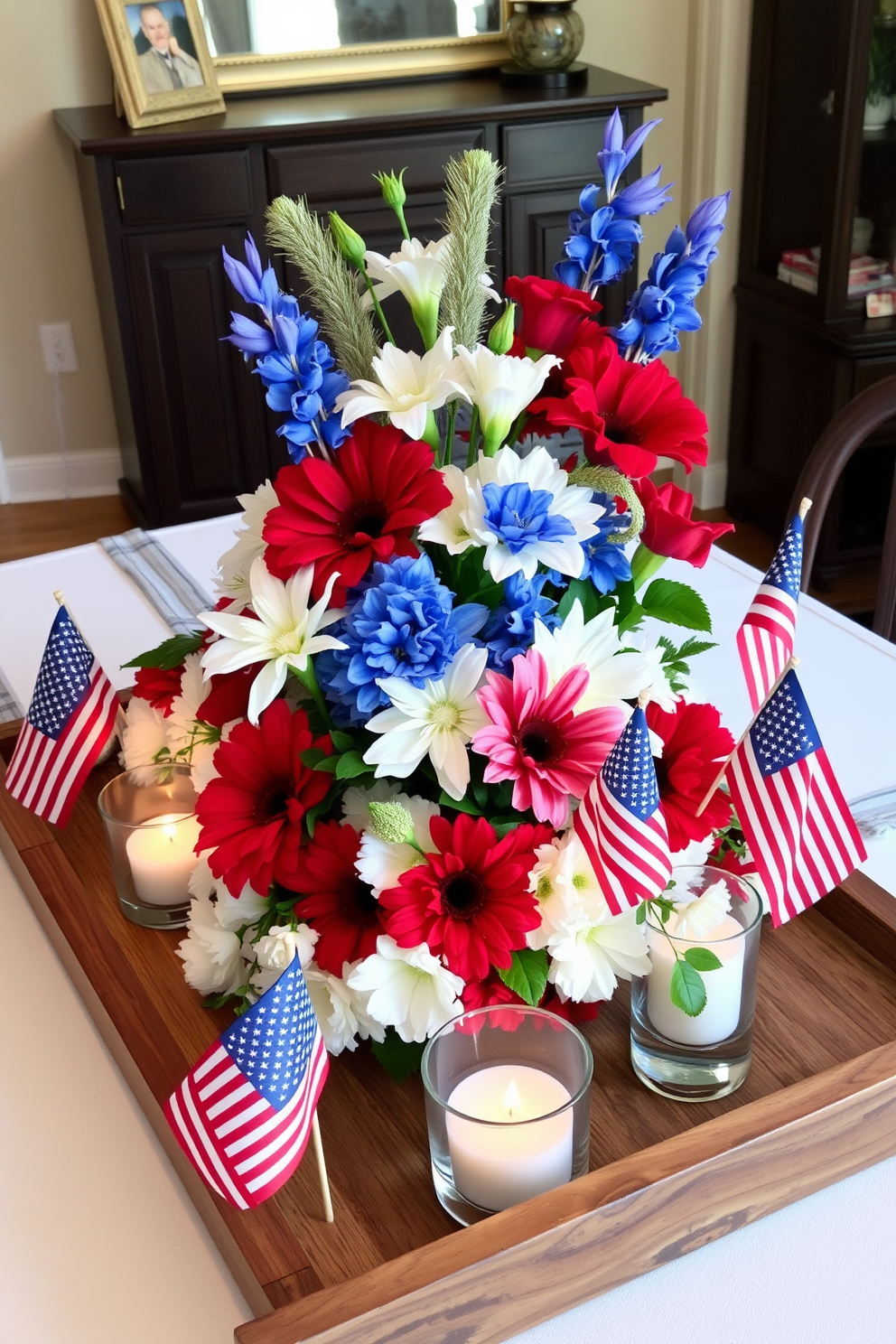 A patriotic themed table centerpiece for Independence Day features a large wooden tray filled with vibrant red white and blue flowers. Surrounding the flowers are small decorative flags and candles in glass holders that create a warm inviting atmosphere.