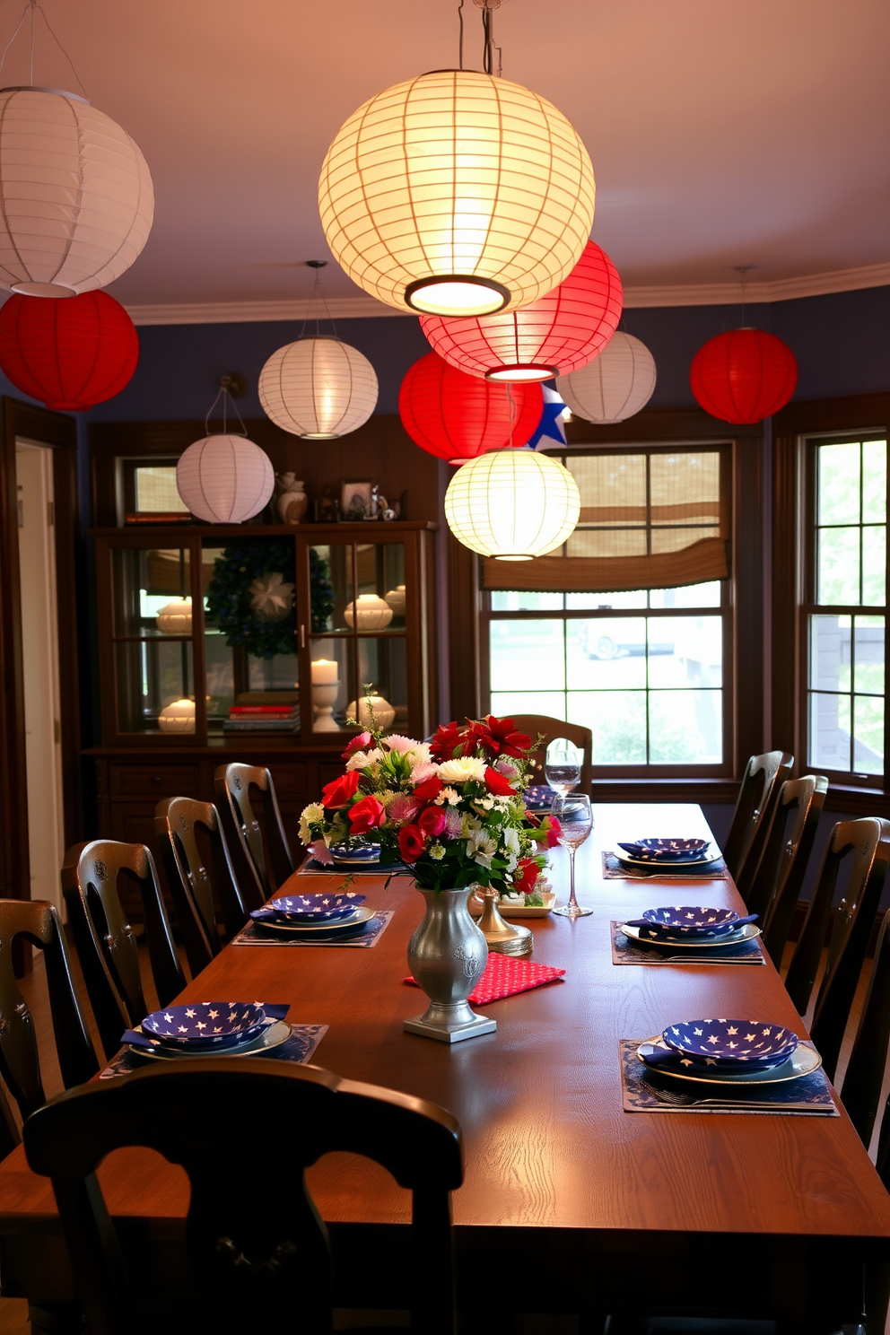 A festive dining room setting adorned with hanging paper lanterns in red white and blue. The lanterns cast a warm glow over a long wooden dining table set with star patterned tableware and fresh flowers in a vintage vase.