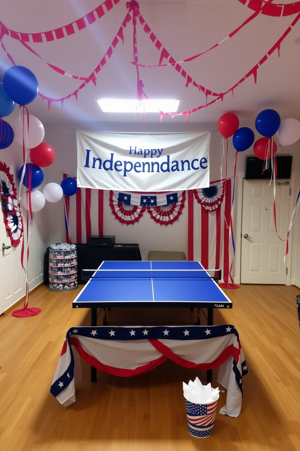 A festive game room setting for the Fourth of July. The table tennis area is adorned with red white and blue decorations including balloons and streamers. A large banner reading Happy Independence Day hangs above the table. The table itself is covered with a patriotic tablecloth and surrounded by themed party supplies.