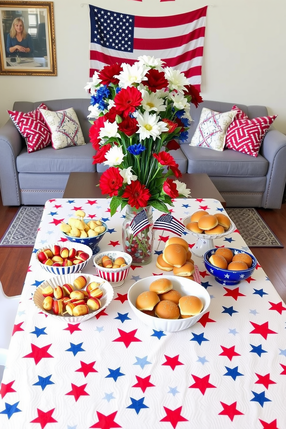 A festive table setting for snacks featuring a red white and blue tablecloth adorned with stars and stripes. On the table, a variety of snacks including fruit skewers and mini burgers are arranged in decorative bowls. The living room is decorated with patriotic accents such as throw pillows and a banner featuring the American flag. A centerpiece of fresh flowers in red white and blue completes the festive atmosphere.