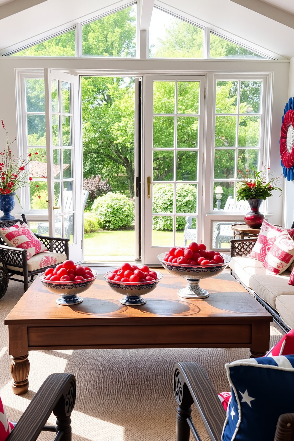 A bright sunroom filled with natural light features a large window that opens to a lush garden. On a wooden coffee table, decorative bowls overflowing with vibrant red fruits create a festive atmosphere for Independence Day celebrations. The walls are adorned with patriotic decor, incorporating shades of red, white, and blue. Comfortable seating with colorful cushions invites guests to relax and enjoy the cheerful ambiance.