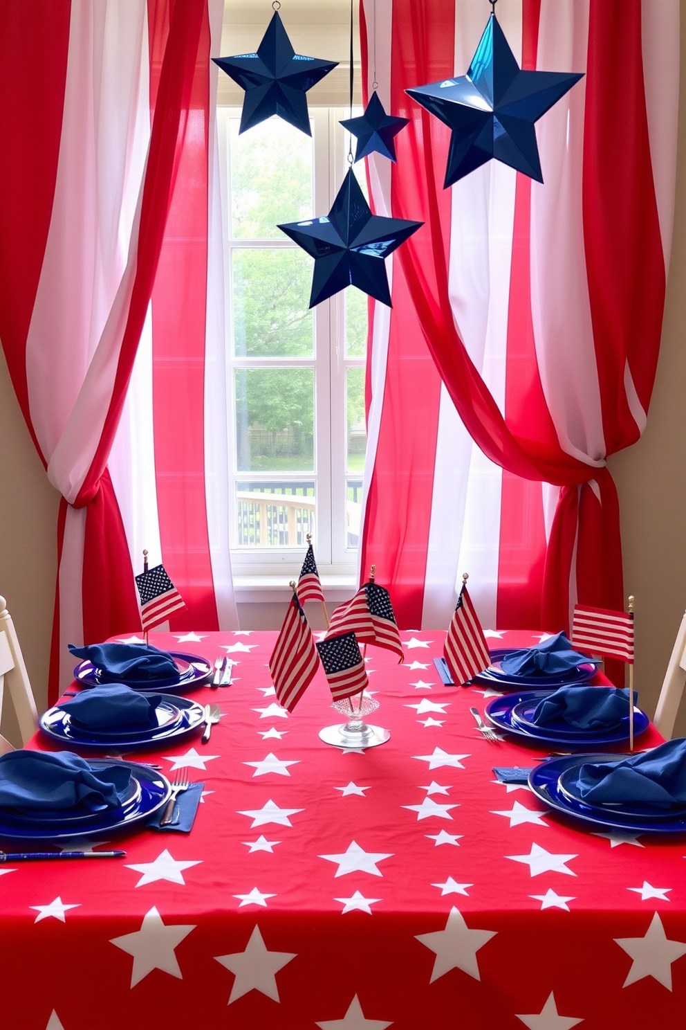 A festive table setting for Independence Day features a vibrant red tablecloth adorned with white star patterns. On top, blue plates are arranged with matching napkins, and small American flags serve as centerpieces. The window is decorated with flowing red and white curtains that frame a view of the outdoors. Hanging from the window are blue star-shaped ornaments that catch the light and add a patriotic touch.