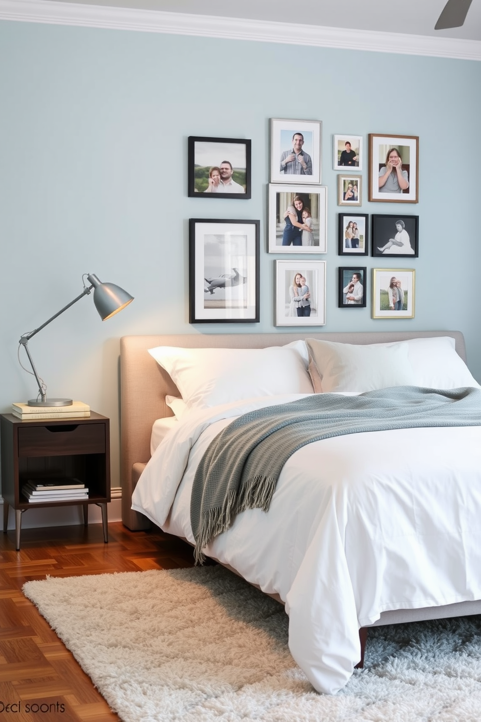 A cozy bedroom featuring a gallery wall adorned with a collection of framed photos showcasing memorable moments. The bed is dressed in crisp white linens with a soft gray throw blanket, creating a serene atmosphere. To the side, a stylish nightstand holds a modern lamp and a stack of books, while a plush area rug adds warmth to the hardwood floor. The walls are painted in a soft blue hue, complementing the artwork displayed on the gallery wall.