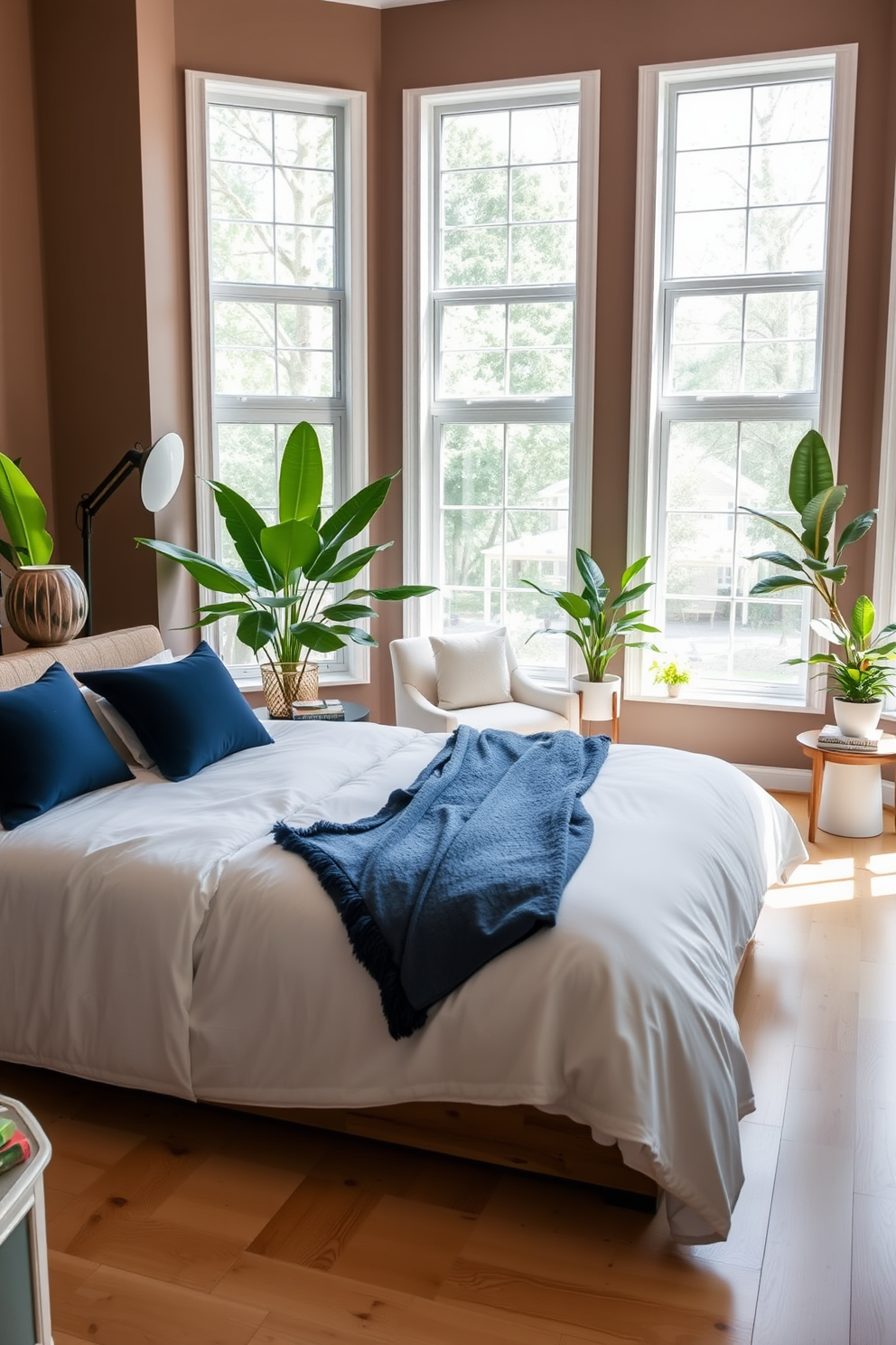 A serene bedroom setting featuring a plush king-size bed dressed in soft white linens and accented with navy blue throw pillows. Large windows allow natural light to flood the room, and potted plants are placed in each corner to create a fresh and inviting atmosphere. The walls are painted in a warm taupe hue, complementing the light hardwood flooring. A stylish bedside table holds a modern lamp and a stack of books, while a cozy reading nook with a comfortable chair and a small side table adds functionality to the space.