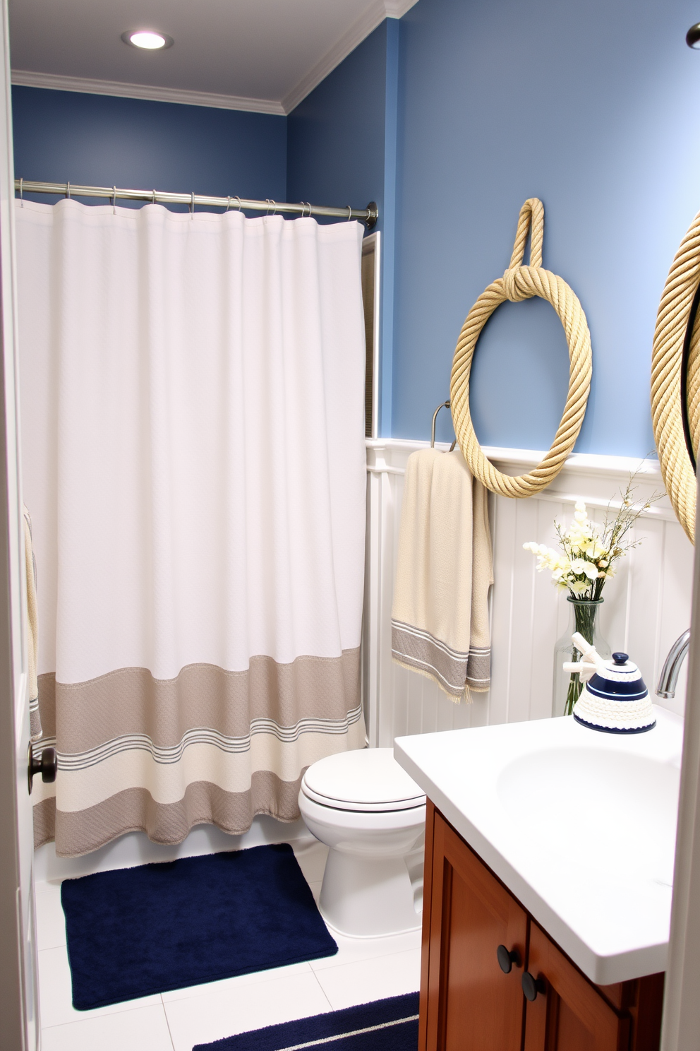 A serene bathroom setting featuring nautical rope accents throughout the decor. The walls are painted in a soft blue hue, complemented by white wainscoting and a wooden vanity adorned with rope handles. Decorative elements include a shower curtain with a subtle nautical pattern and a matching bath mat. Accents like a rope-wrapped mirror and a vase filled with seashells complete the Memorial Day theme.