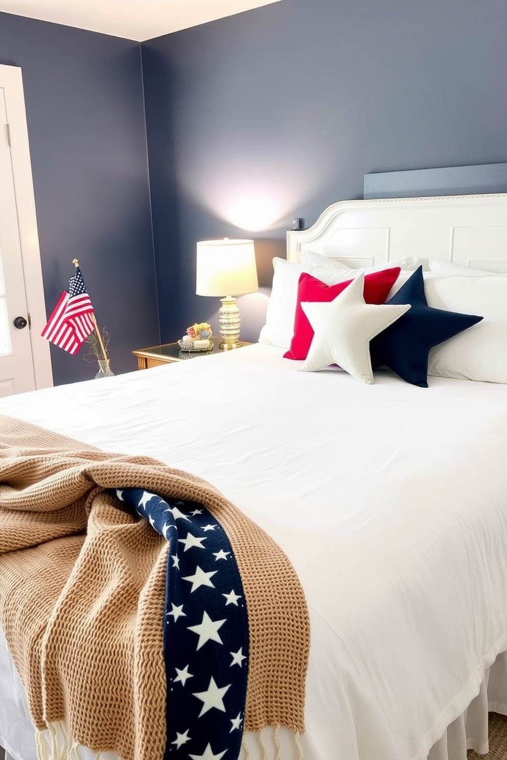 A cozy bedroom featuring a crisp white bedspread adorned with star shaped decorative pillows in shades of red, white, and blue. The walls are painted a soft navy blue, and a vintage wooden nightstand holds a small American flag in a glass vase. A festive touch is added with a woven throw blanket draped over the foot of the bed, complementing the star shaped pillows. Soft lighting from a bedside lamp creates a warm and inviting atmosphere, perfect for celebrating Memorial Day.