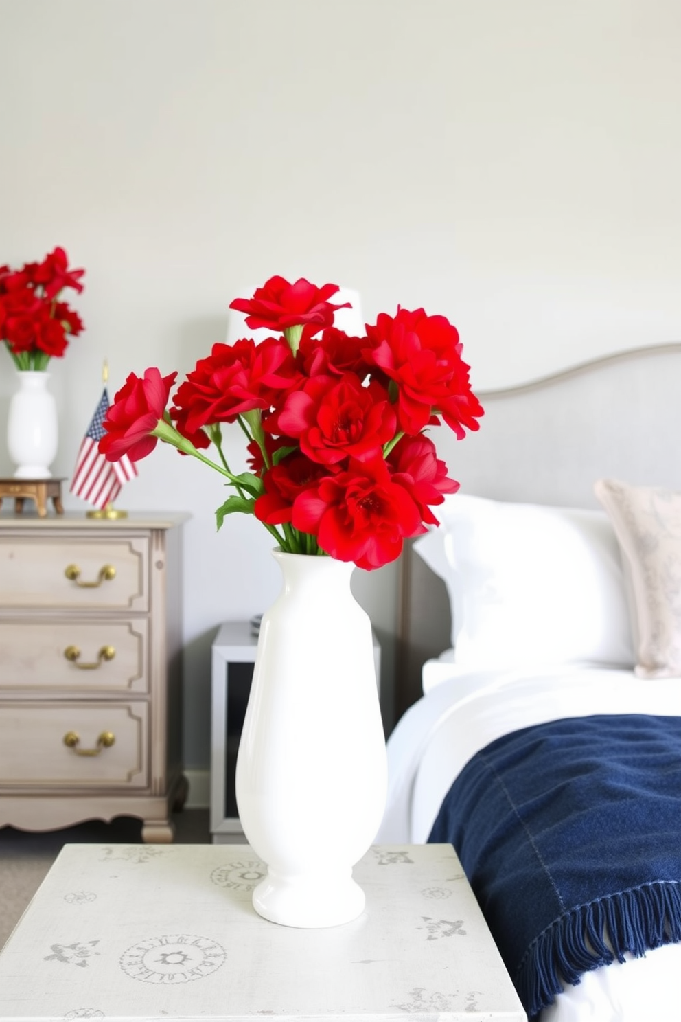 A serene bedroom setting featuring decorative vases filled with vibrant red flowers. The vases are placed on a bedside table next to a plush bed adorned with crisp white linens and a navy blue throw blanket. The walls are painted in a soft gray hue, creating a calming atmosphere. A vintage dresser with brass handles complements the decor, and a small American flag is elegantly displayed on the dresser in honor of Memorial Day.