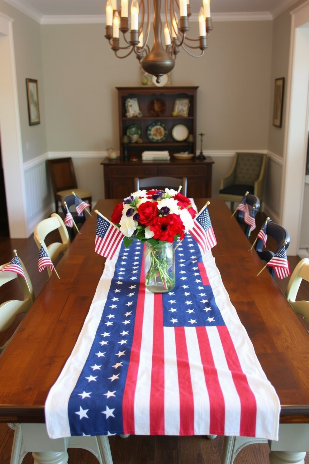 A patriotic table runner featuring stars and stripes is elegantly draped across a long wooden dining table. Surrounding the table are mismatched vintage chairs, each adorned with small American flags for a festive touch. The center of the table showcases a vibrant bouquet of red, white, and blue flowers in a rustic mason jar. Soft, warm lighting from a chandelier above creates a welcoming atmosphere for a Memorial Day gathering.