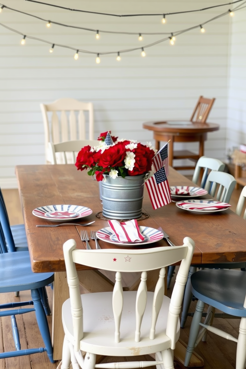 Themed dinnerware featuring stars and stripes is set on a rustic wooden dining table. Surrounding the table are mismatched chairs painted in shades of blue and white, creating a casual yet festive atmosphere. A centerpiece of red and white flowers is arranged in a vintage metal bucket. String lights are draped above, casting a warm glow over the space, enhancing the celebratory vibe for Memorial Day.