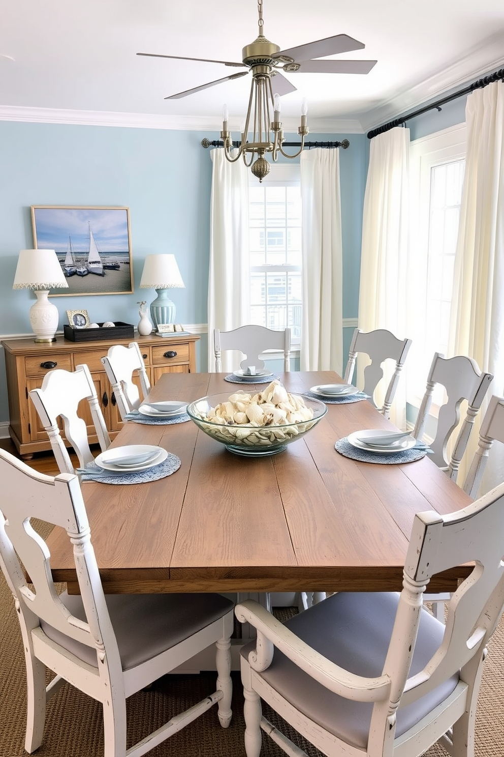 A nautical themed dining room features a large wooden table with a weathered finish surrounded by whitewashed chairs. Seashell accents are tastefully incorporated into the table setting with a centerpiece of assorted seashells arranged in a glass bowl. The walls are painted in a soft blue hue reminiscent of the ocean, while nautical-themed artwork adorns the walls. Light, airy curtains flutter in the breeze, completing the coastal vibe of this Memorial Day dining room.