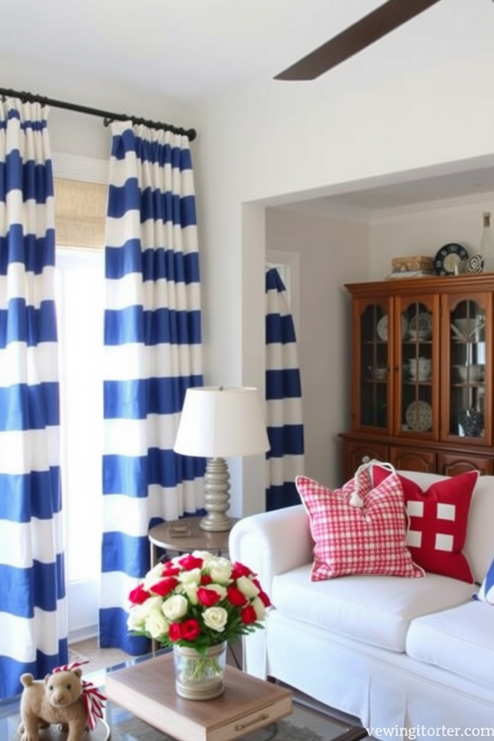 A bright and airy living room features curtains adorned with nautical stripes in shades of blue and white. The space is accented with red and white decorative pillows, creating a festive atmosphere for Memorial Day celebrations.