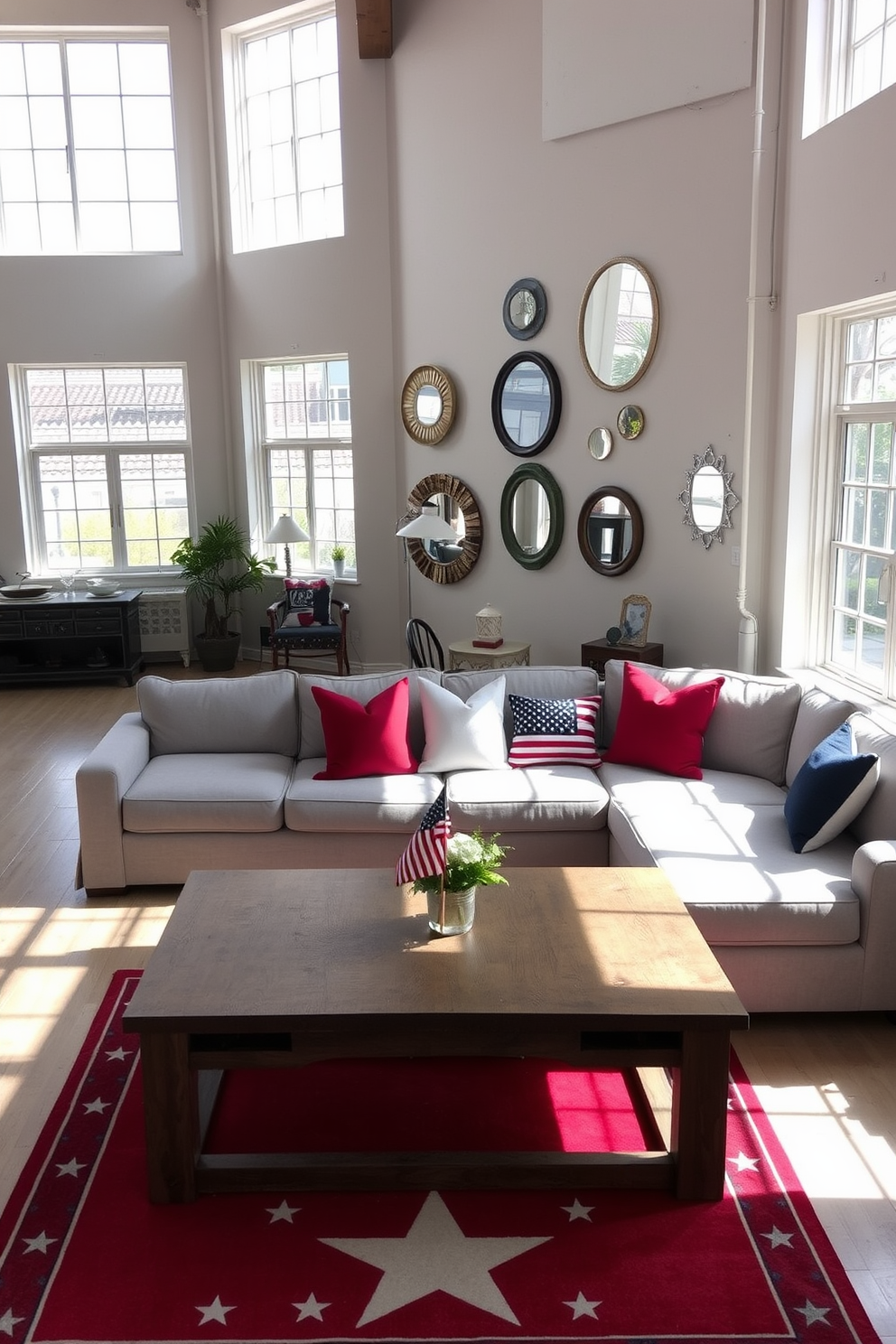 A spacious loft living area with large windows that flood the space with natural light. The walls are painted in a soft white hue, and a collection of decorative mirrors is strategically placed to reflect the sunlight throughout the room. The furnishings include a plush sectional sofa in a light gray fabric and a rustic wooden coffee table at the center. To celebrate Memorial Day, red, white, and blue throw pillows are arranged on the sofa, complemented by a vibrant area rug featuring a subtle star pattern.