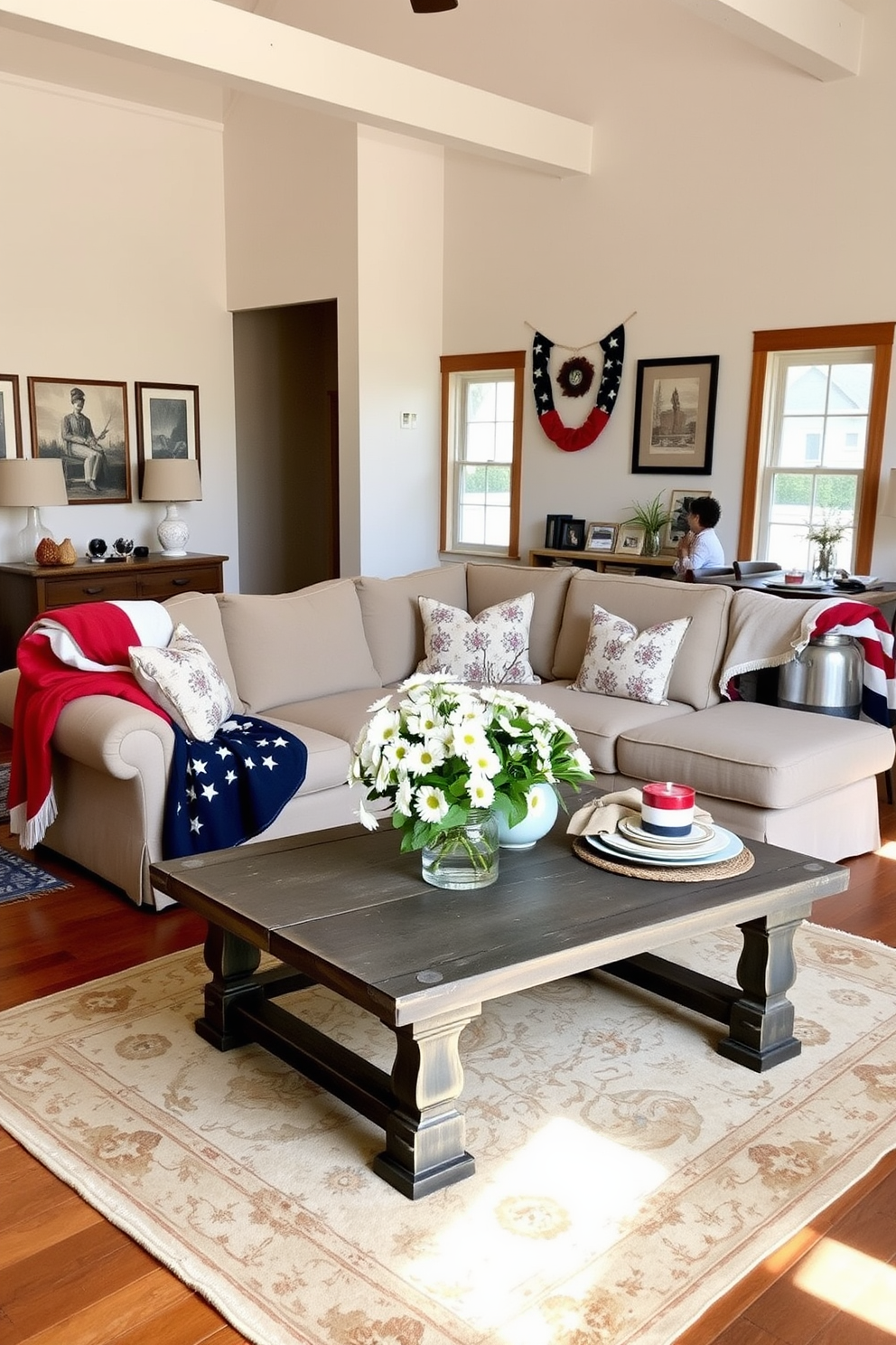 A bright and airy loft space adorned with seasonal decor celebrating Memorial Day. The room features a large, comfortable sectional sofa draped with red, white, and blue throw blankets, and a rustic coffee table topped with a floral arrangement of fresh daisies and patriotic accents. On the walls, framed artwork reflects the spirit of the season, while a vintage-inspired rug adds warmth to the wooden floor. A small dining area is set with a festive tablecloth and decorative place settings, creating an inviting atmosphere for gatherings.