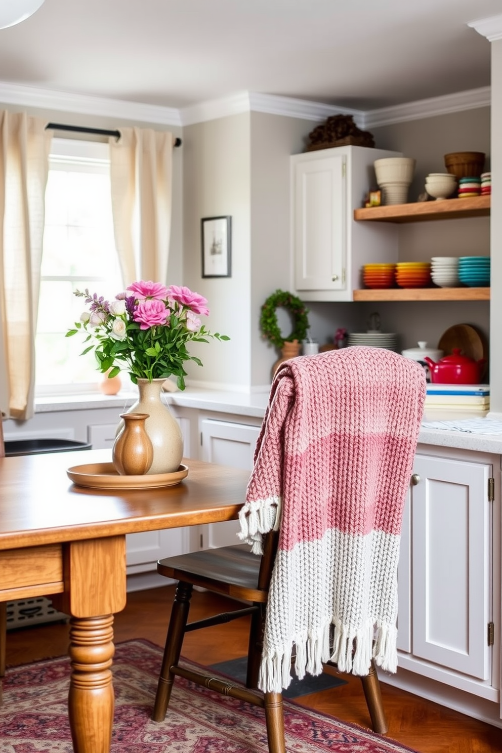 A warm and inviting kitchen filled with layered textiles that create a cozy atmosphere. Soft linen curtains frame the windows while a chunky knit throw is draped over a wooden chair, enhancing the space's charm. The countertops are adorned with fresh flowers in a ceramic vase and colorful dishware arranged artfully. A patterned area rug lies beneath the dining table, adding texture and warmth to the overall design.