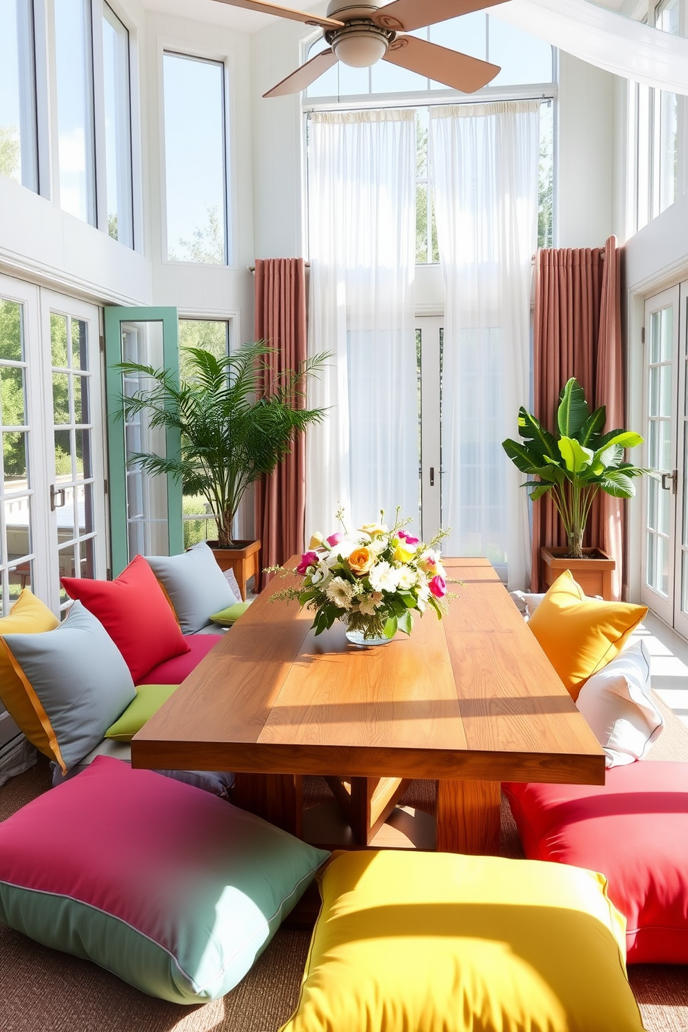 A bright and airy summer dining room features a large wooden table surrounded by oversized cushions in vibrant colors for casual seating. Natural light floods the space through large windows, illuminating the fresh floral centerpiece and the light, breezy curtains that flutter gently in the breeze.