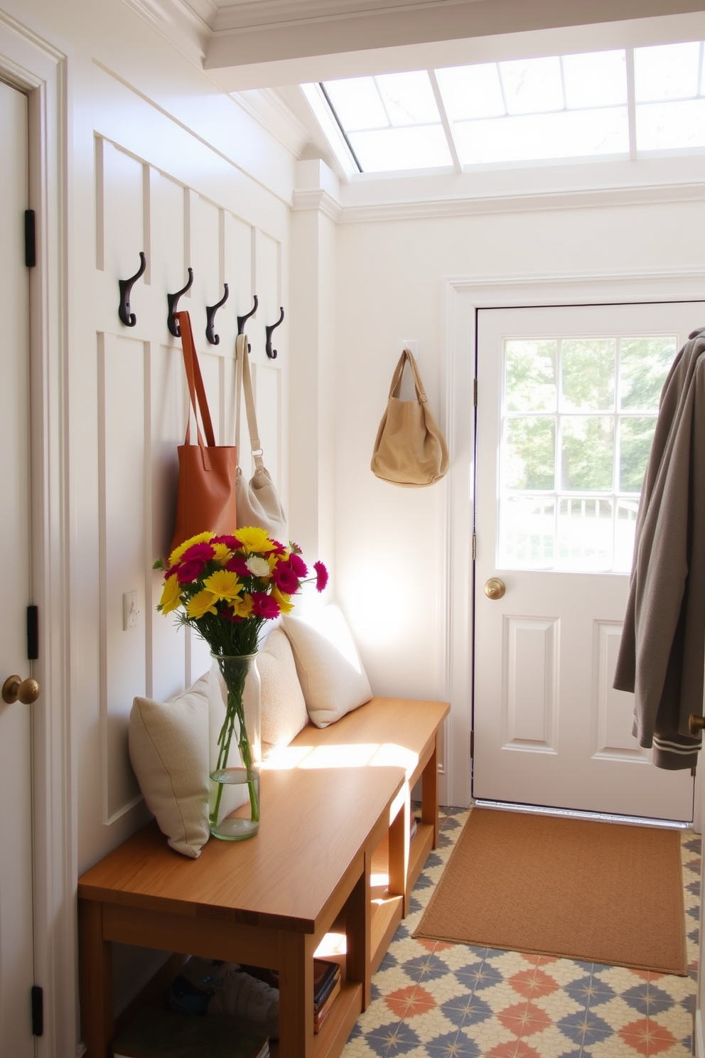 A bright and airy mudroom filled with natural light. The space features a wooden bench with plush cushions and hooks for hanging bags and coats. Fresh flowers in a vibrant vase sit on the bench, adding a cheerful touch. The walls are painted in a soft white, while the floor is adorned with colorful patterned tiles.