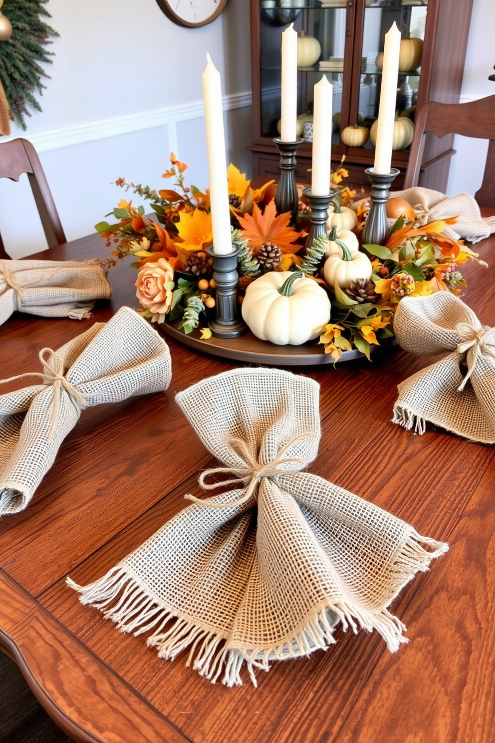 Burlap napkin holders with twine accents are elegantly arranged on a rustic wooden dining table. The table is adorned with a warm, autumnal centerpiece featuring seasonal fruits, candles, and small pumpkins for a cozy Thanksgiving atmosphere.