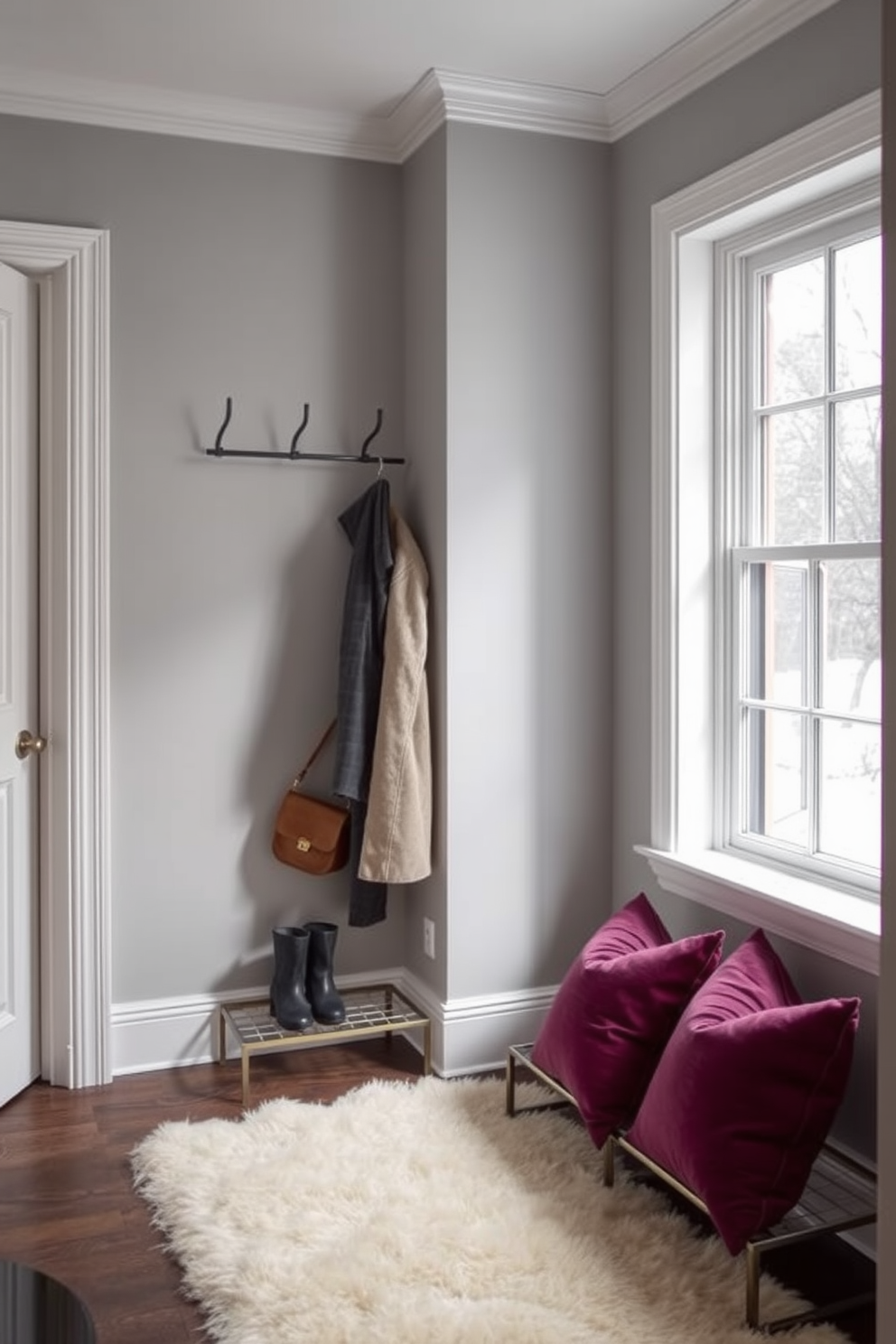 A stylish winter closet features a chic coat rack made of sleek metal with a minimalist design. The walls are adorned with soft gray paint, and the floor is covered in a plush cream rug for added warmth. The coat rack is elegantly positioned near a large window, allowing natural light to illuminate the space. A decorative bench sits beneath the window, complemented by cozy throw pillows in rich jewel tones.