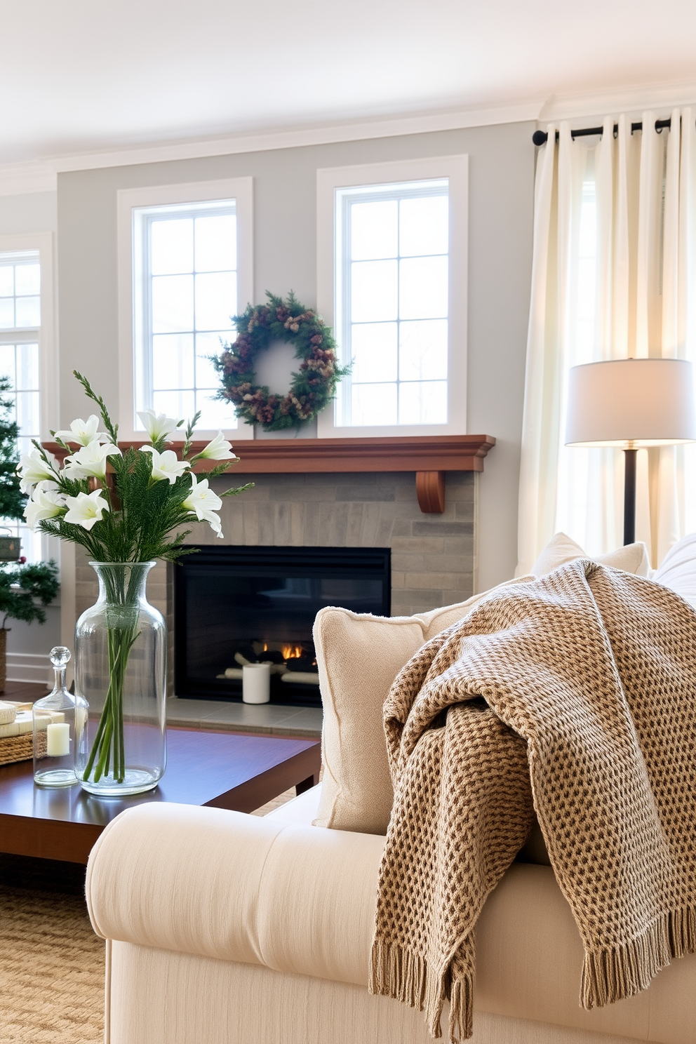 A cozy winter living room filled with natural light. The focal point is a large fireplace adorned with glass vases filled with seasonal winter florals like white lilies and evergreen sprigs. Soft, plush furniture in neutral tones invites relaxation. A woven throw blanket drapes over the arm of a cream-colored sofa, complementing the warm ambiance of the space.